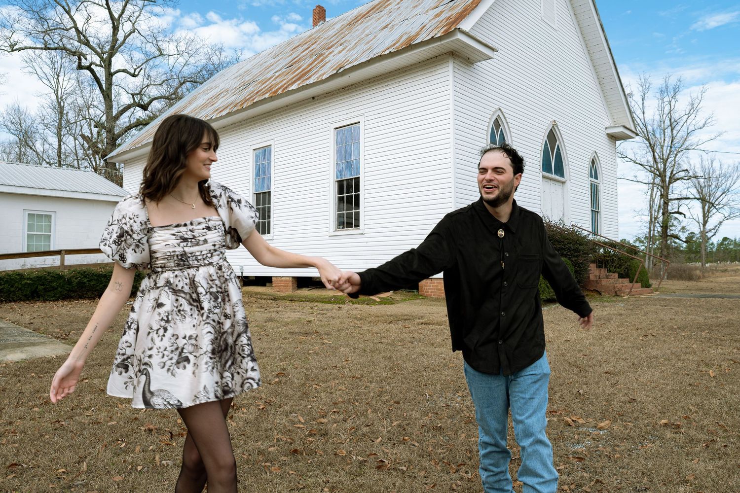 Couple holding hands and smiling outside a white country church on a winter day.