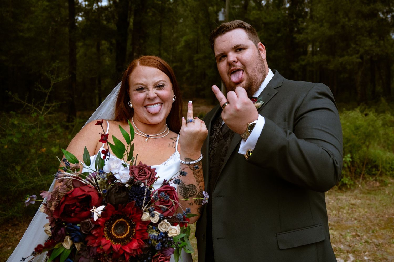 A wedding couple poses playfully in the woods with a burgundy and white floral bouquet.