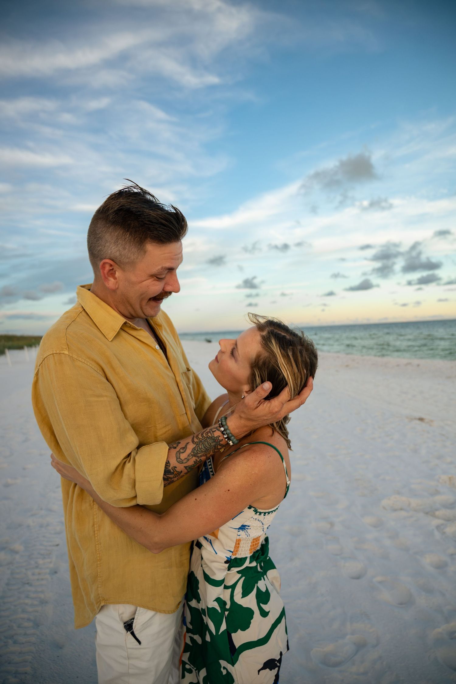 Couple embracing romantically on a white sand beach at sunset, man in yellow shirt holding woman's face tenderly.