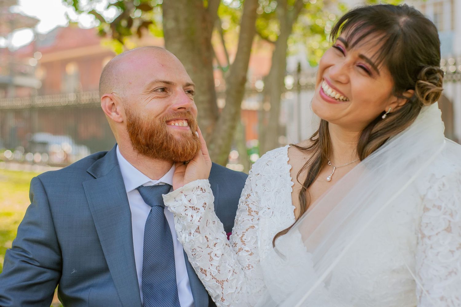 A joyful bride in white lace dress laughs alongside groom in blue suit during outdoor wedding photoshoot.
