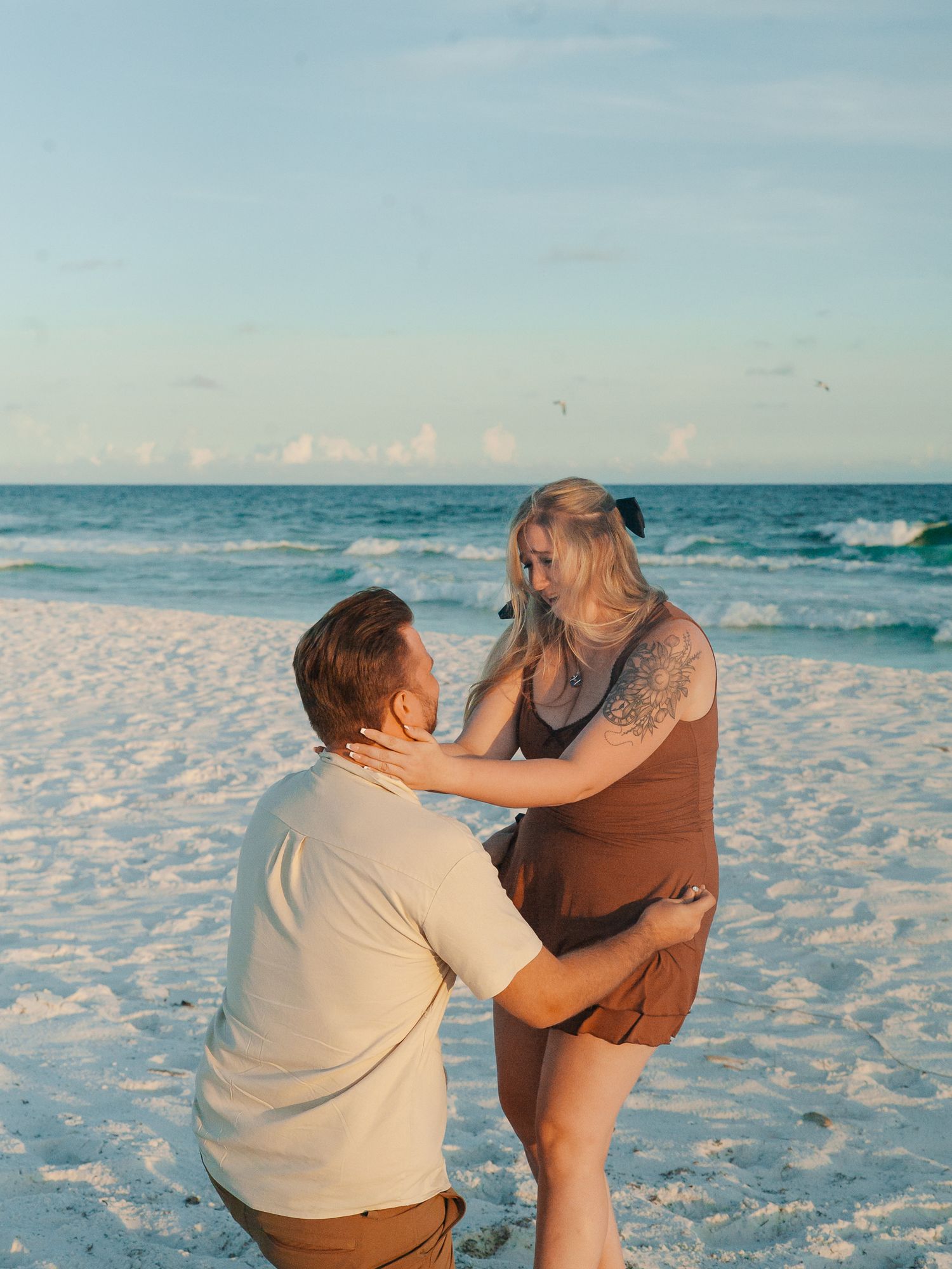 A romantic couple plays and laughs together while sitting on white sandy beach during sunset by the ocean waves.