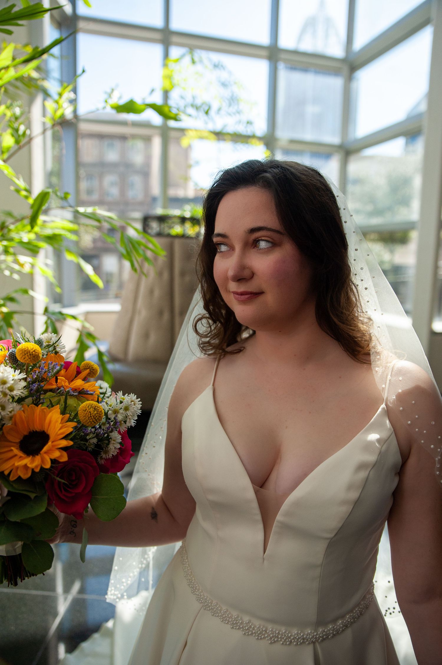 Wedding dress detail shots showing elegant white gown with plunging neckline and crystal belt in sunlit conservatory setting.