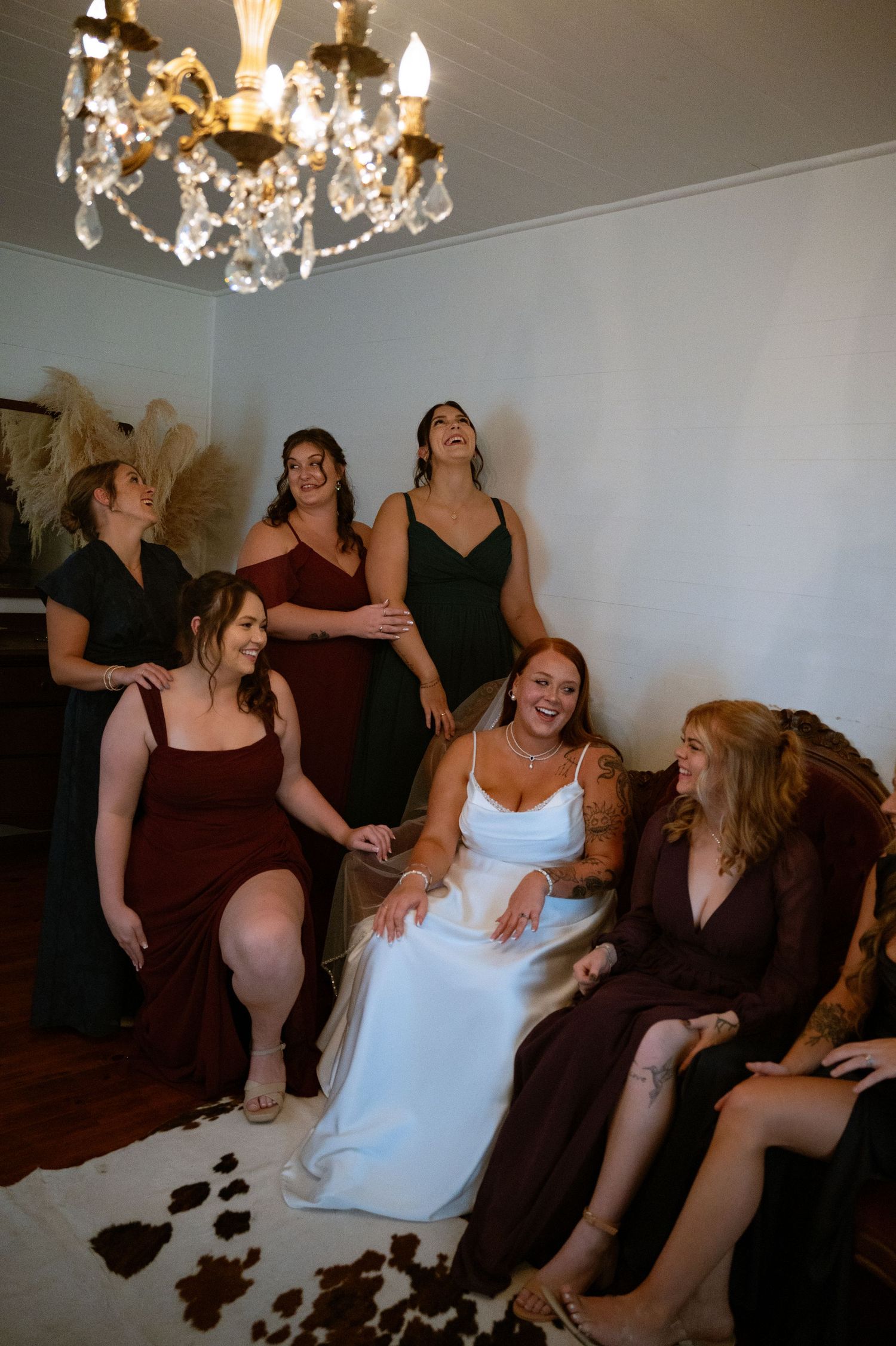 A group in burgundy dresses and one in white sit together laughing and enjoying a celebratory moment under chandeliers.