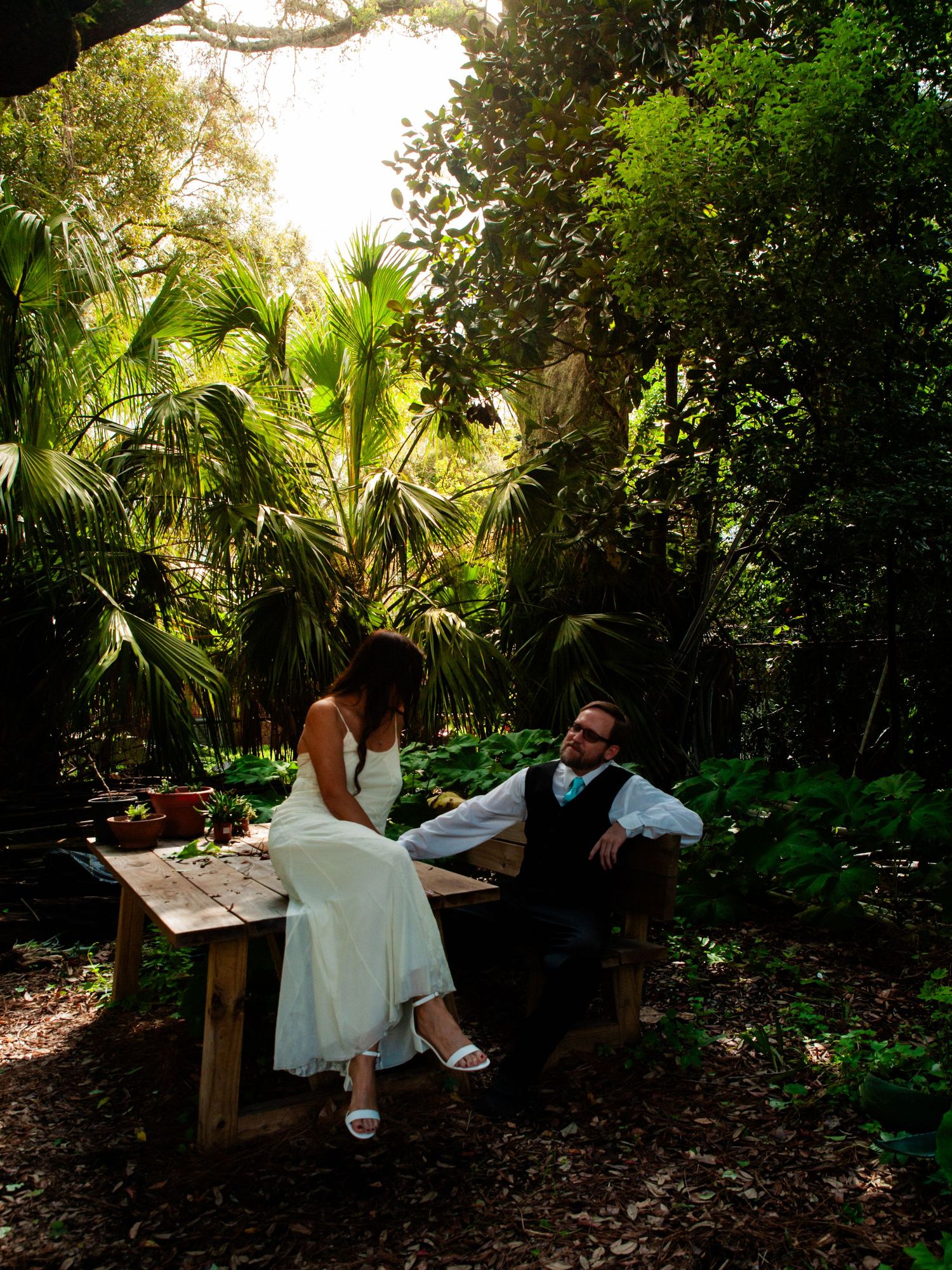 A couple relaxes together on a wooden bench surrounded by lush tropical foliage in a serene garden setting.