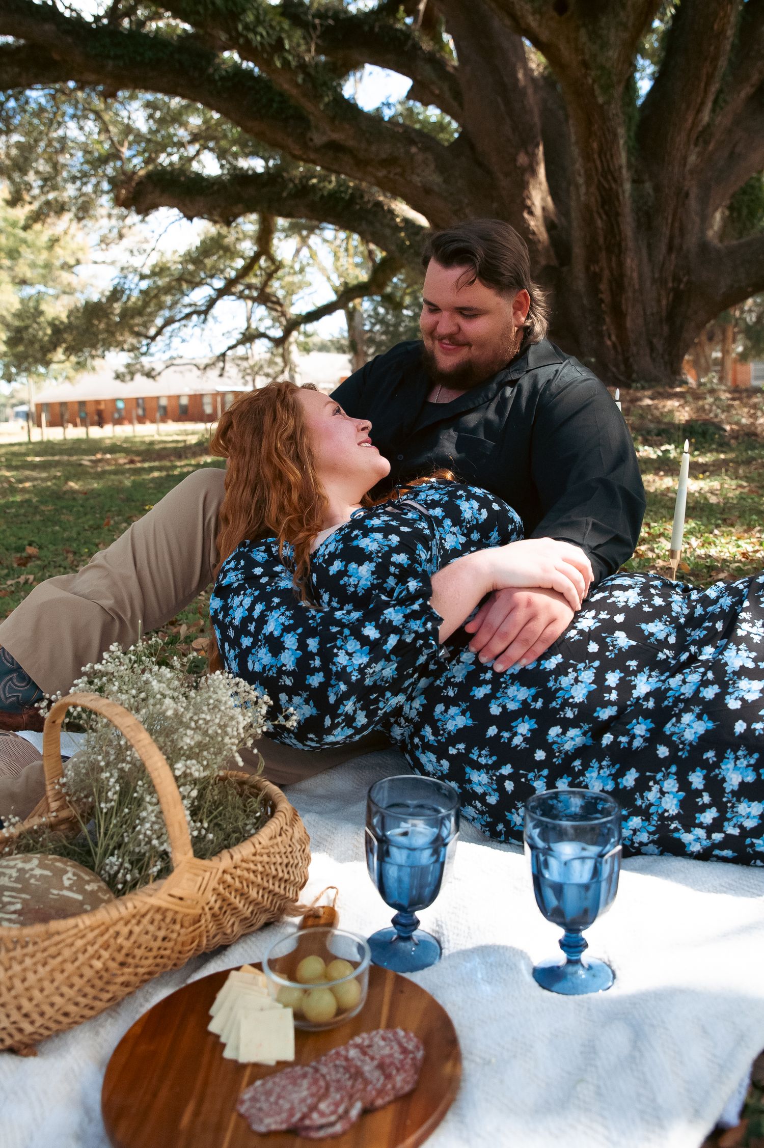 A couple relaxes on a picnic blanket with blue glasses and charcuterie.