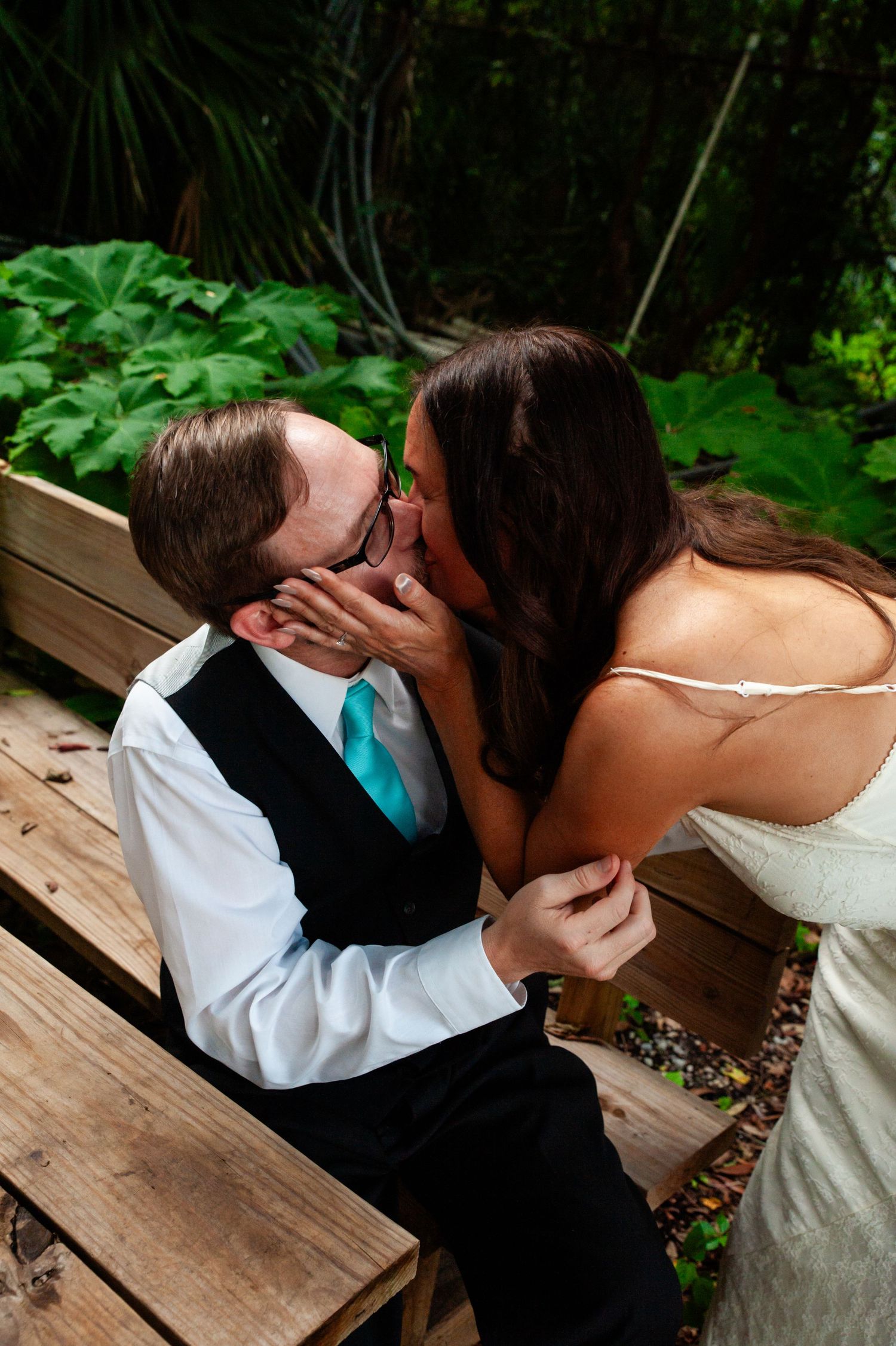 A romantic kiss shared between a couple on wooden decking surrounded by lush greenery at a wedding celebration.