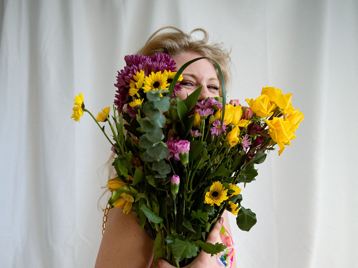 A vibrant bouquet of sunflowers, daffodils and purple blooms held against a white curtain backdrop.