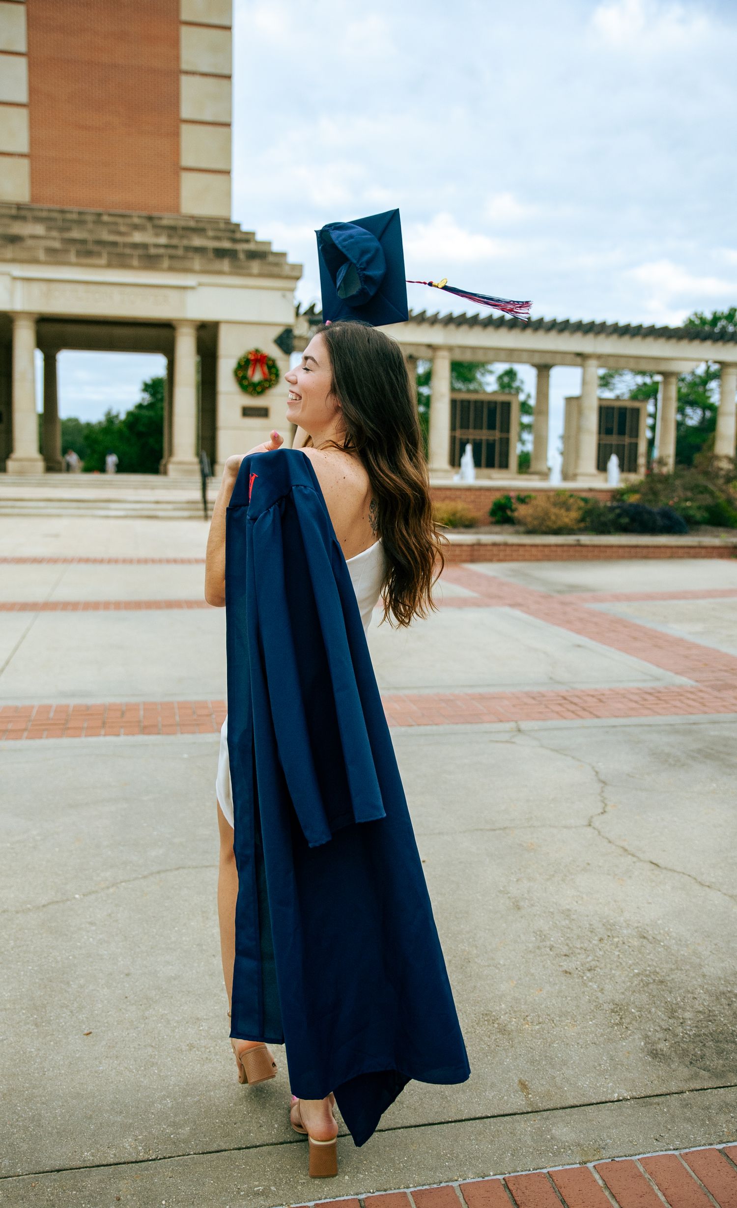 Graduate in navy blue cap and gown poses for graduation photos outside a brick building on a cloudy day.