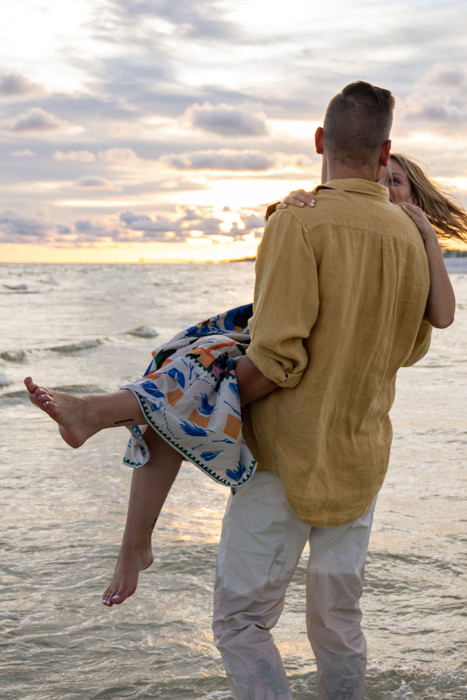 A couple embraces at sunset on the beach, man carrying woman in colorful skirt by the ocean waves.