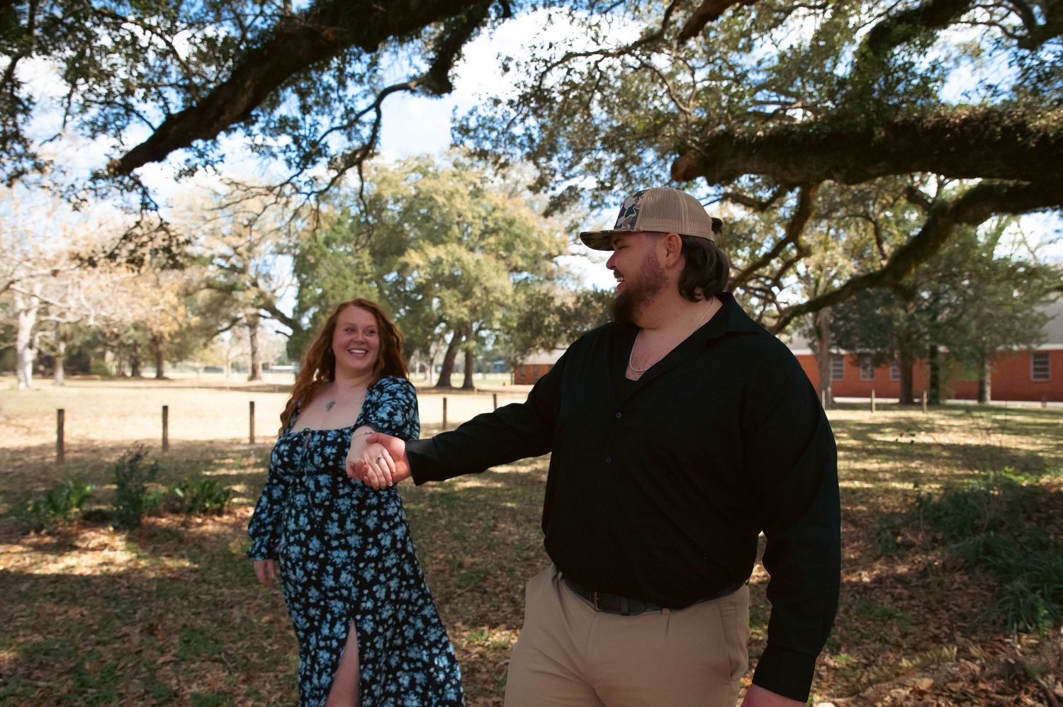 A romantic couple embraces under a large oak tree on a sunny afternoon.