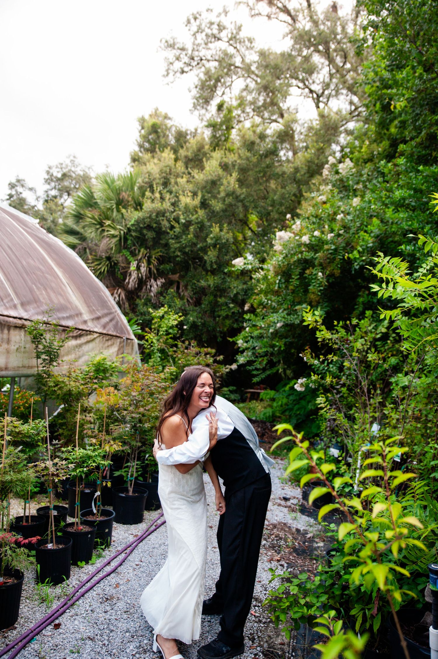A bride in a white dress and dark jacket enjoys a romantic moment in a garden greenhouse setting.