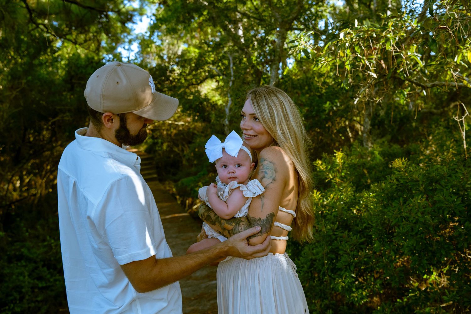 A couple in white outfits shares tender moments while holding their baby in a lush outdoor garden setting.