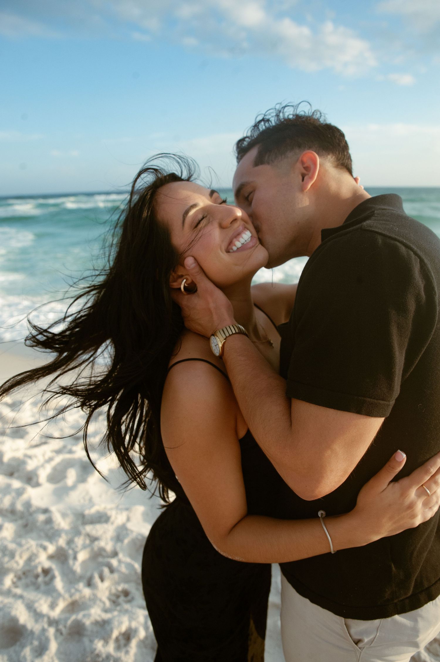 A romantic couple embraces and kisses on a sunny beach with ocean waves in the background.