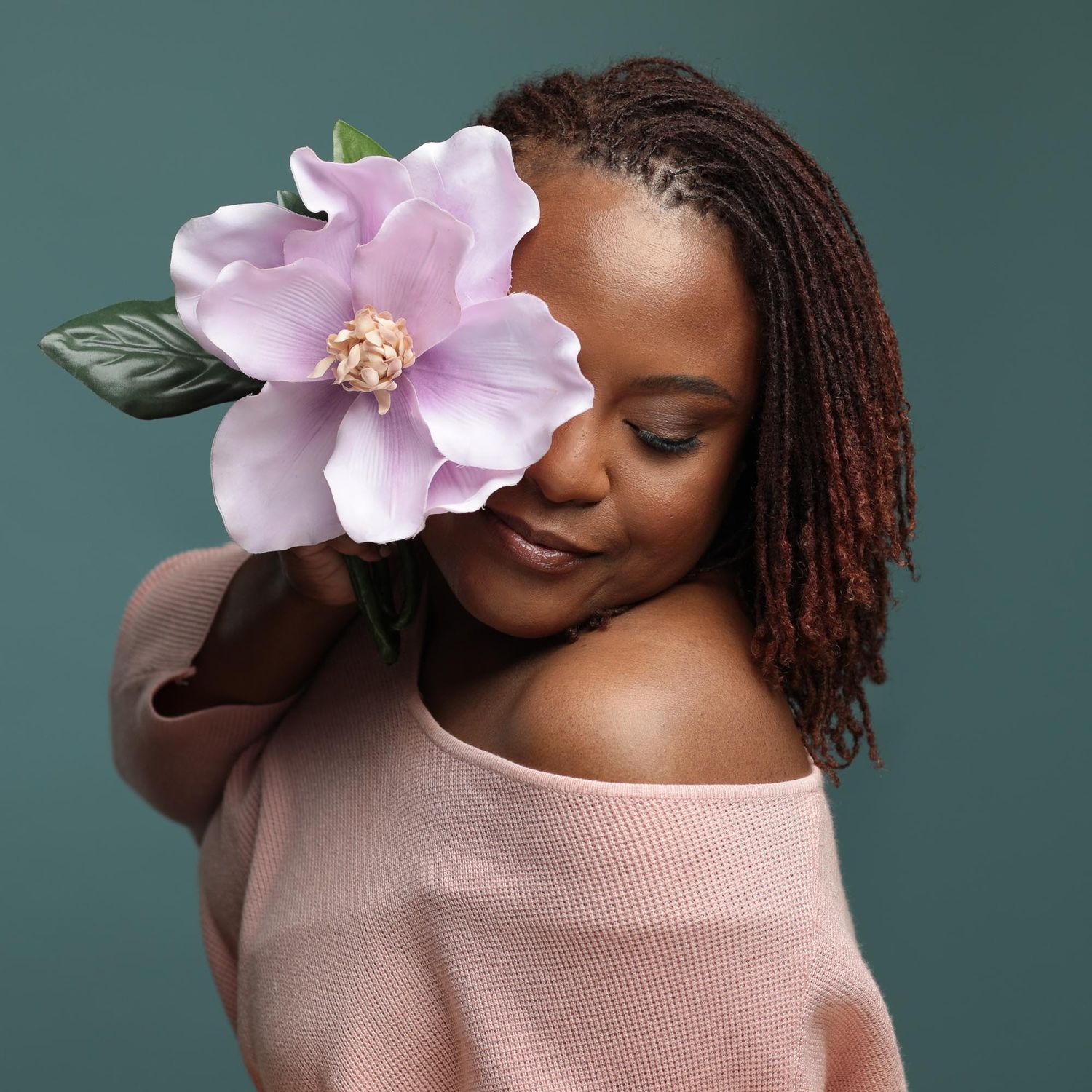 Delicate pink flower decorates a soft portrait against teal background while wearing an off-shoulder pink sweater.