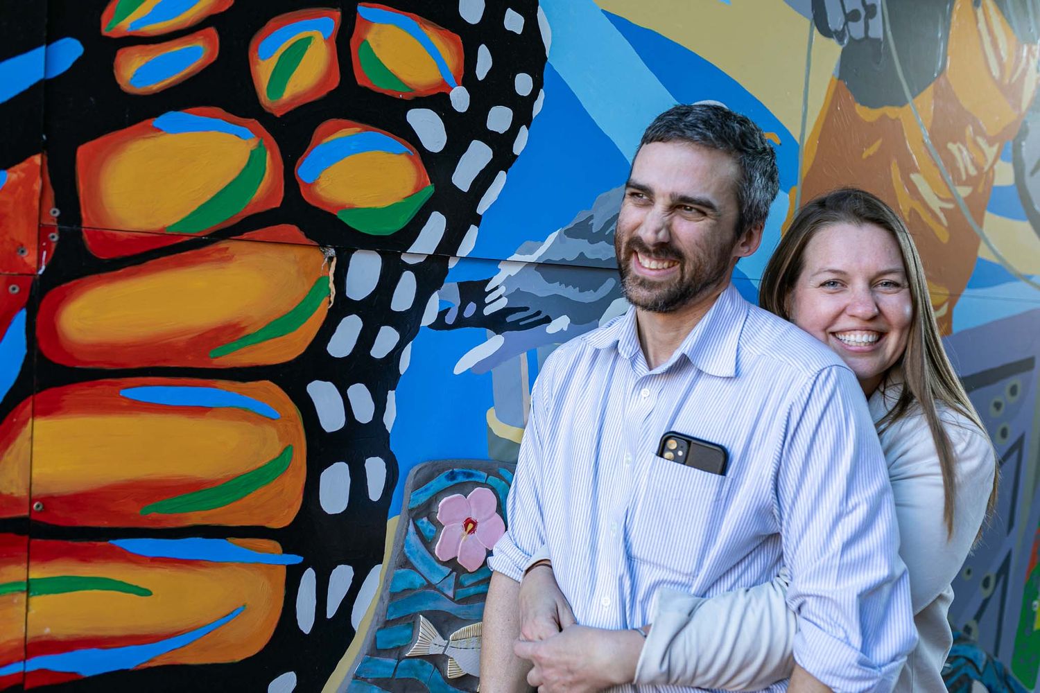 Two people smile while standing in front of a vibrant butterfly wing mural with bright orange and blue colors.