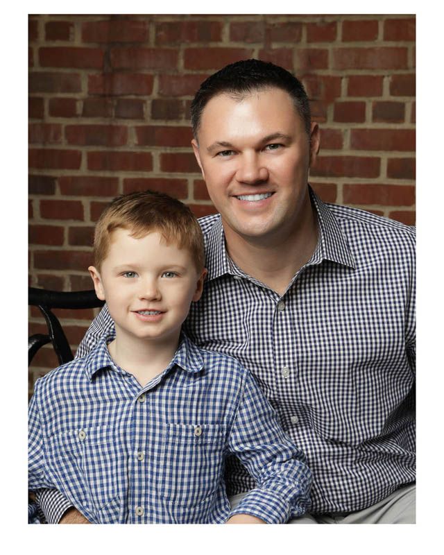 Parent and child in matching navy gingham shirts sitting together and smiling at camera against brick wall.