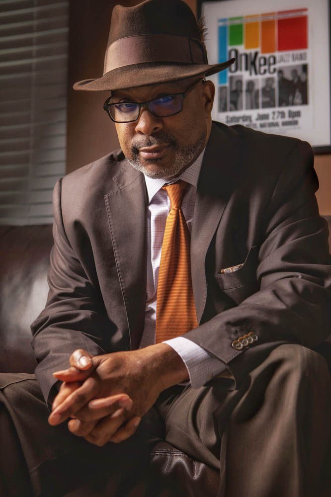 A person in a brown suit and orange tie sits thoughtfully against a dark background.