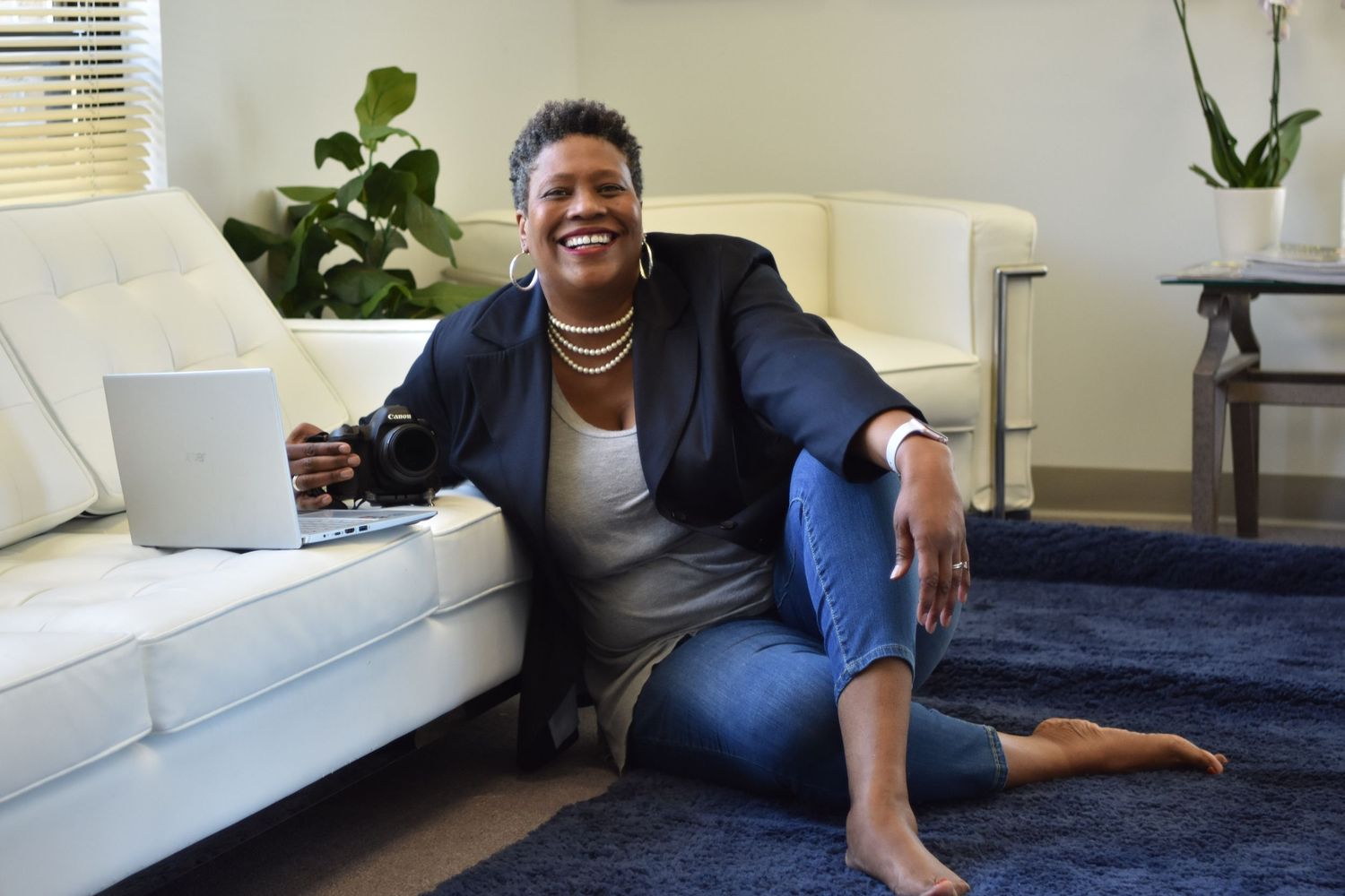 African American woman sits comfortably on the floor in a bright room with white decor, smiling at the camera.
