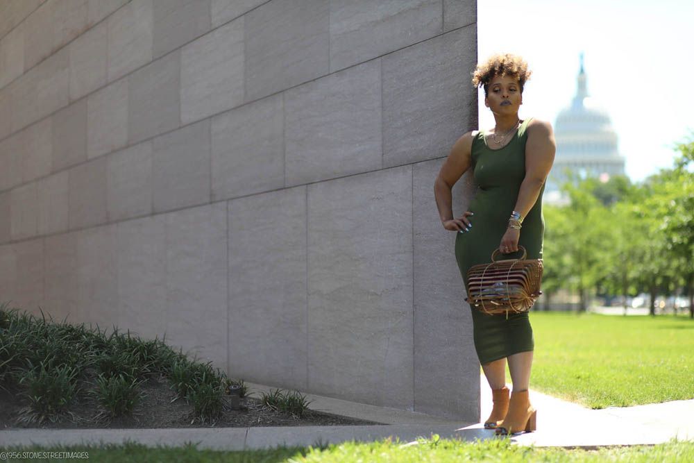 A person in a green dress stands against a concrete wall with the Capitol building visible in the distance.