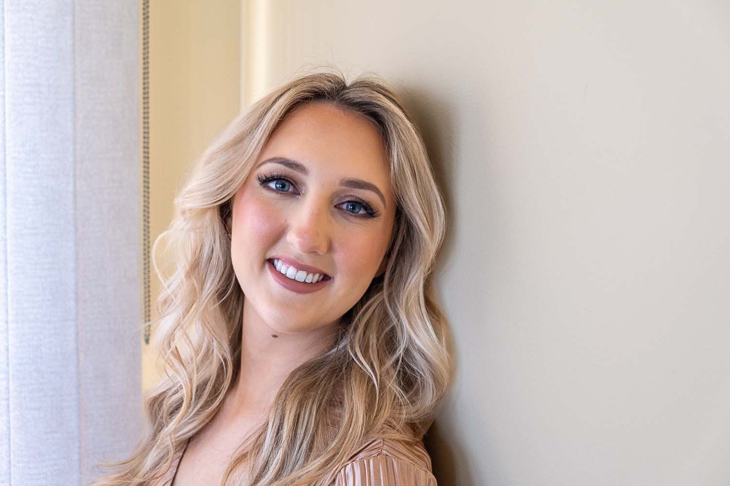 A person with long wavy blonde hair wearing a pink top smiles warmly while posing against a neutral wall.