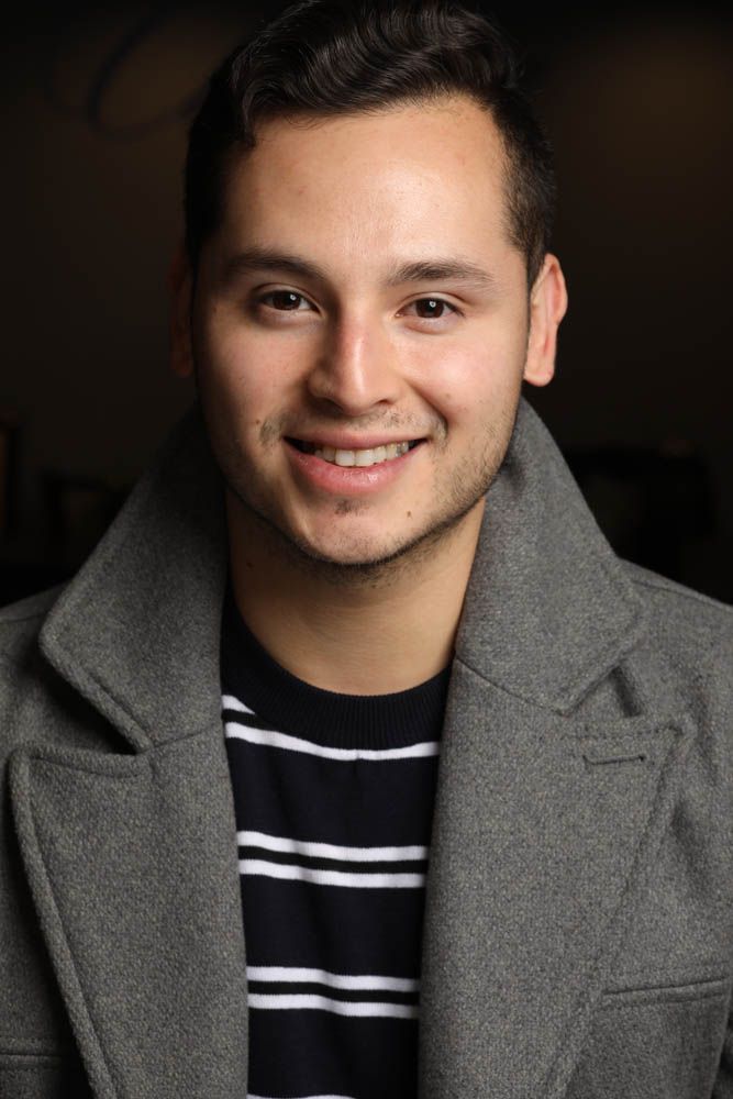 Person in gray coat and striped shirt smiling against dark background in professional headshot series.
