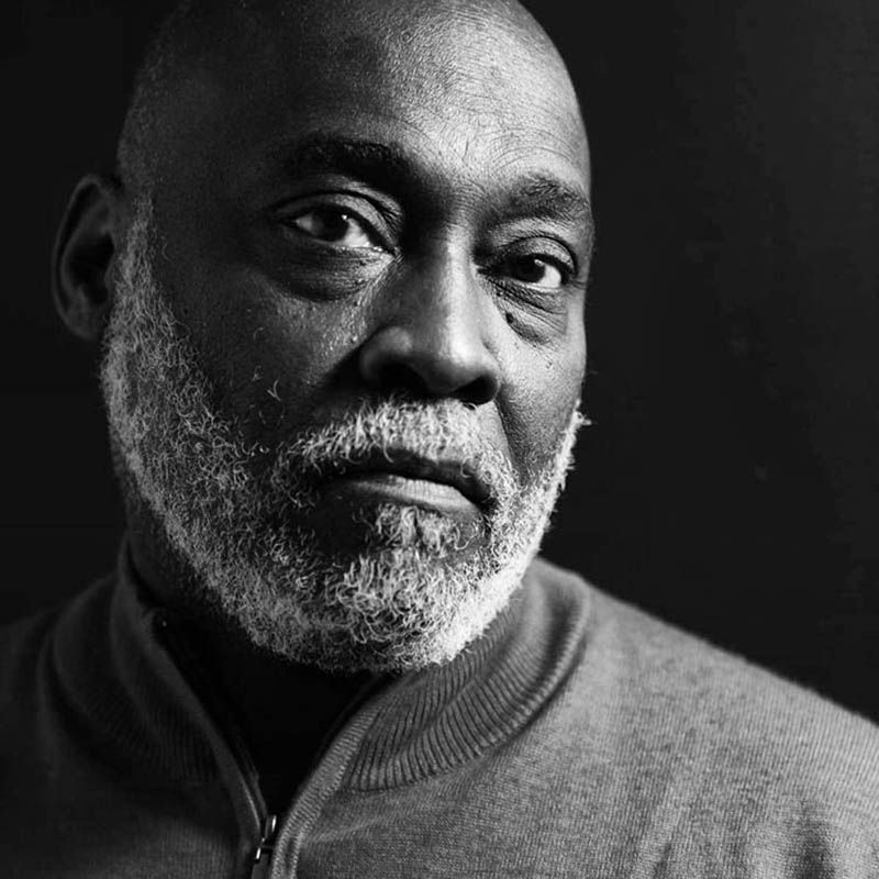 Dramatic black and white close-up portrait showing an older man with white beard and gentle expression against dark background.