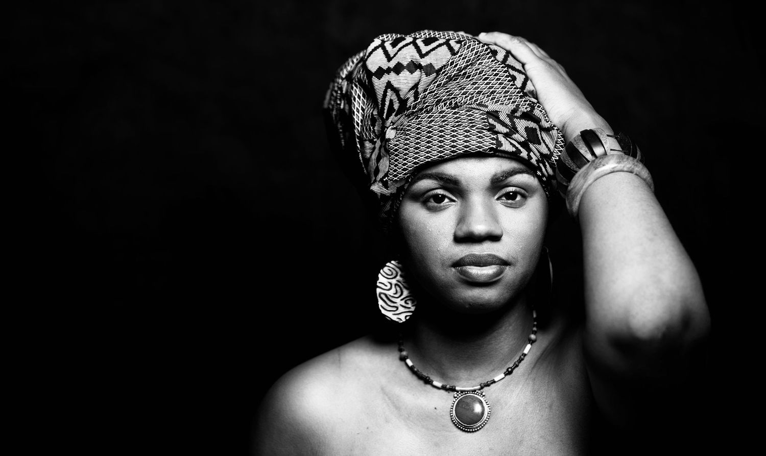 Black and white artistic portrait featuring decorative head wrap and statement necklace against dark background.