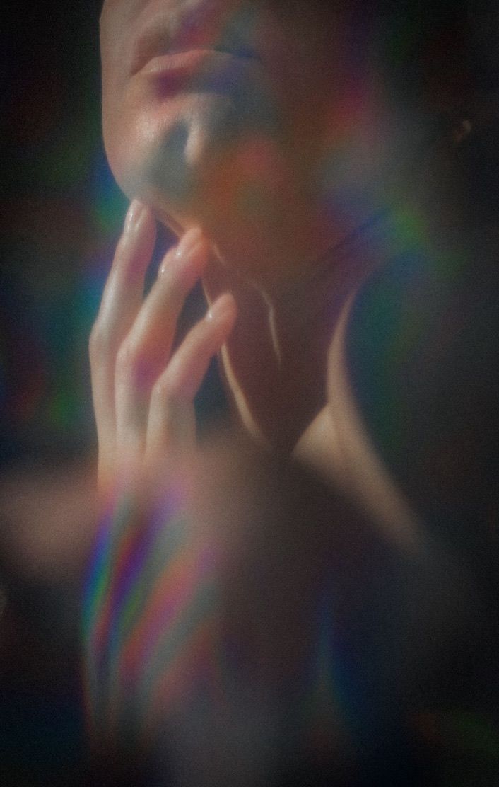 Artistic portrait with ethereal rainbow prism effects showing hands touching face in soft focus dreamy lighting.