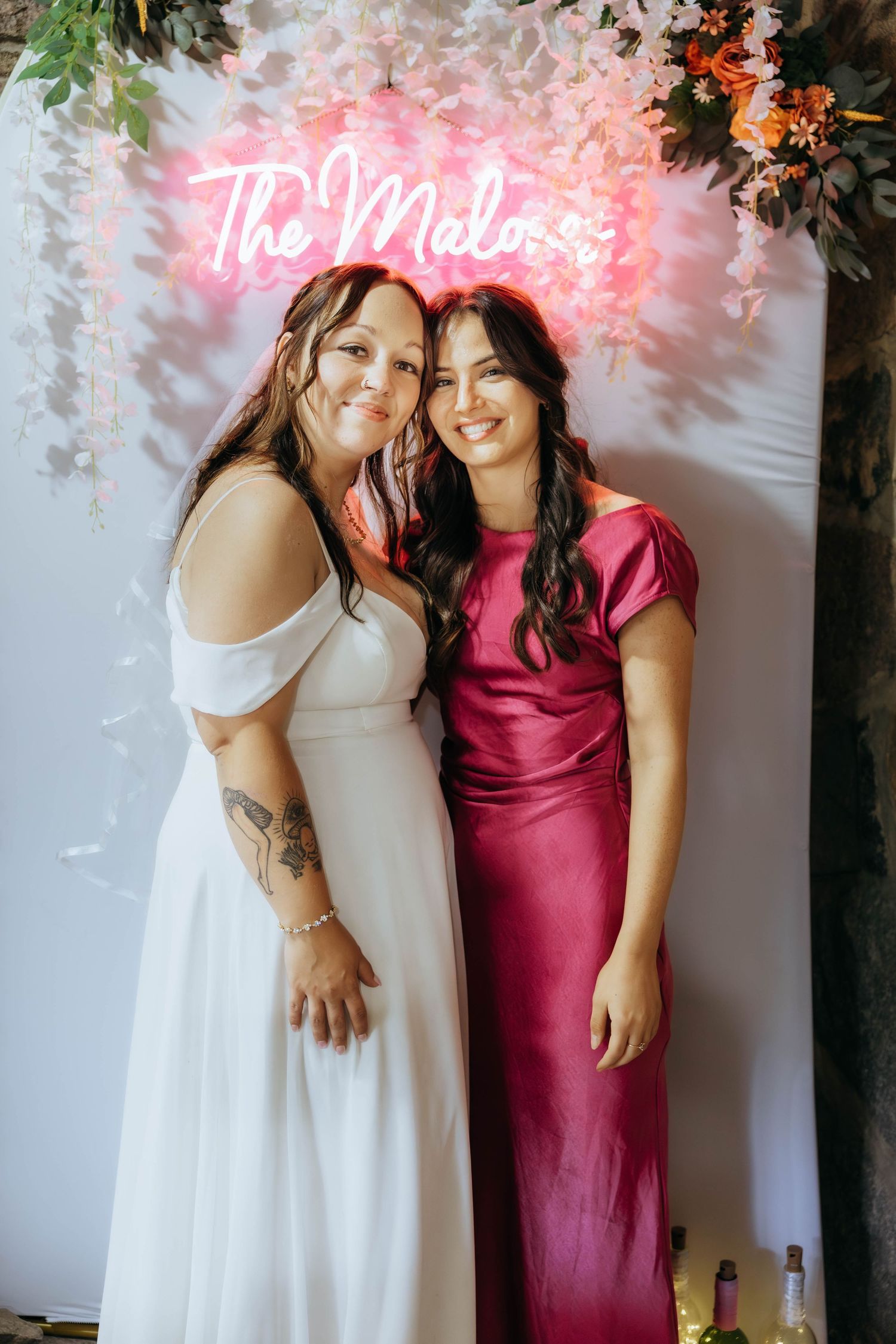 Wedding party members posing together against floral wall with pink neon sign.