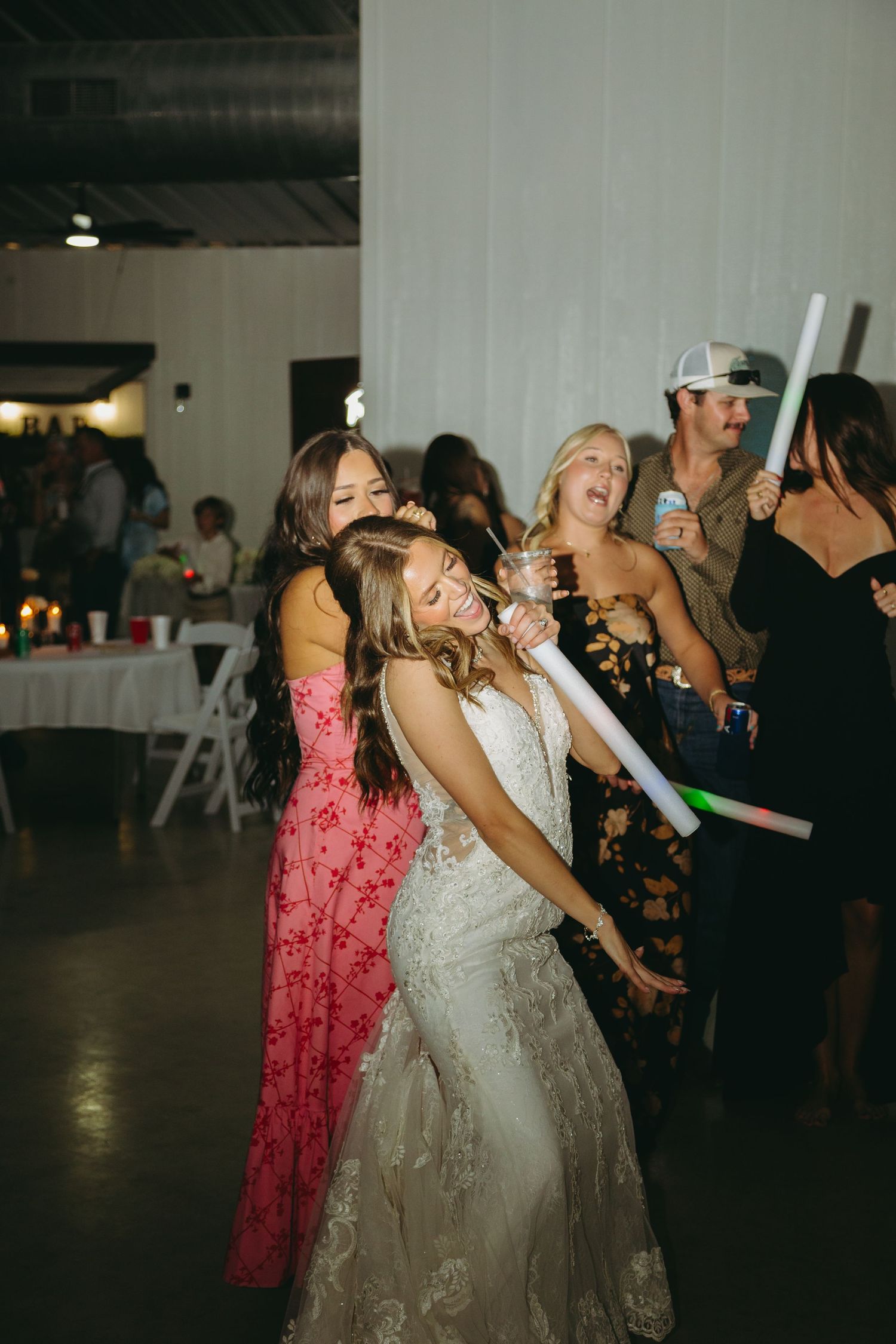 Wedding celebration with guests dancing and holding glowing foam sticks in a dimly lit venue.