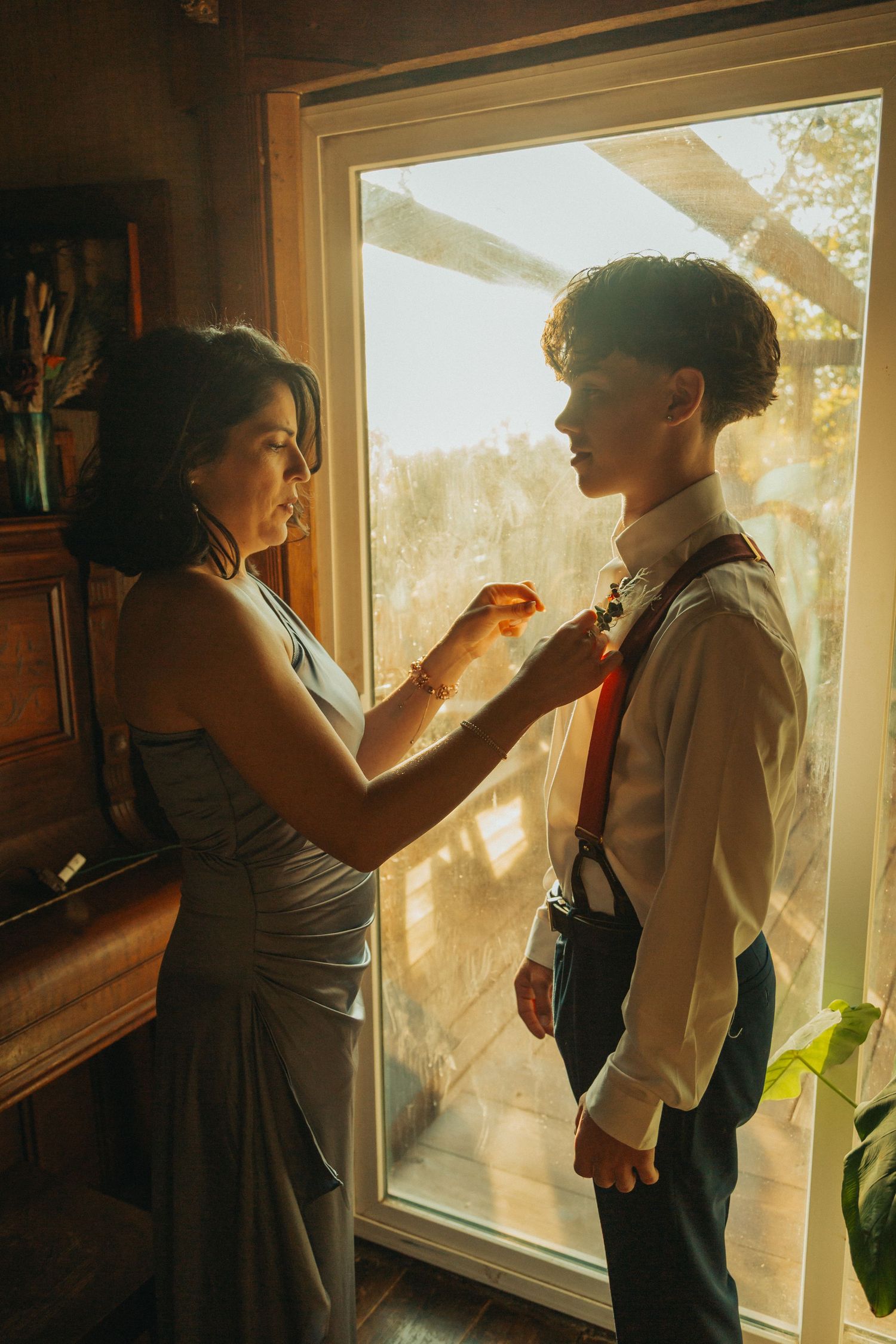 Sunlit window illuminates a scene of a woman placing a boutonnière on a young man before a wedding ceremony.