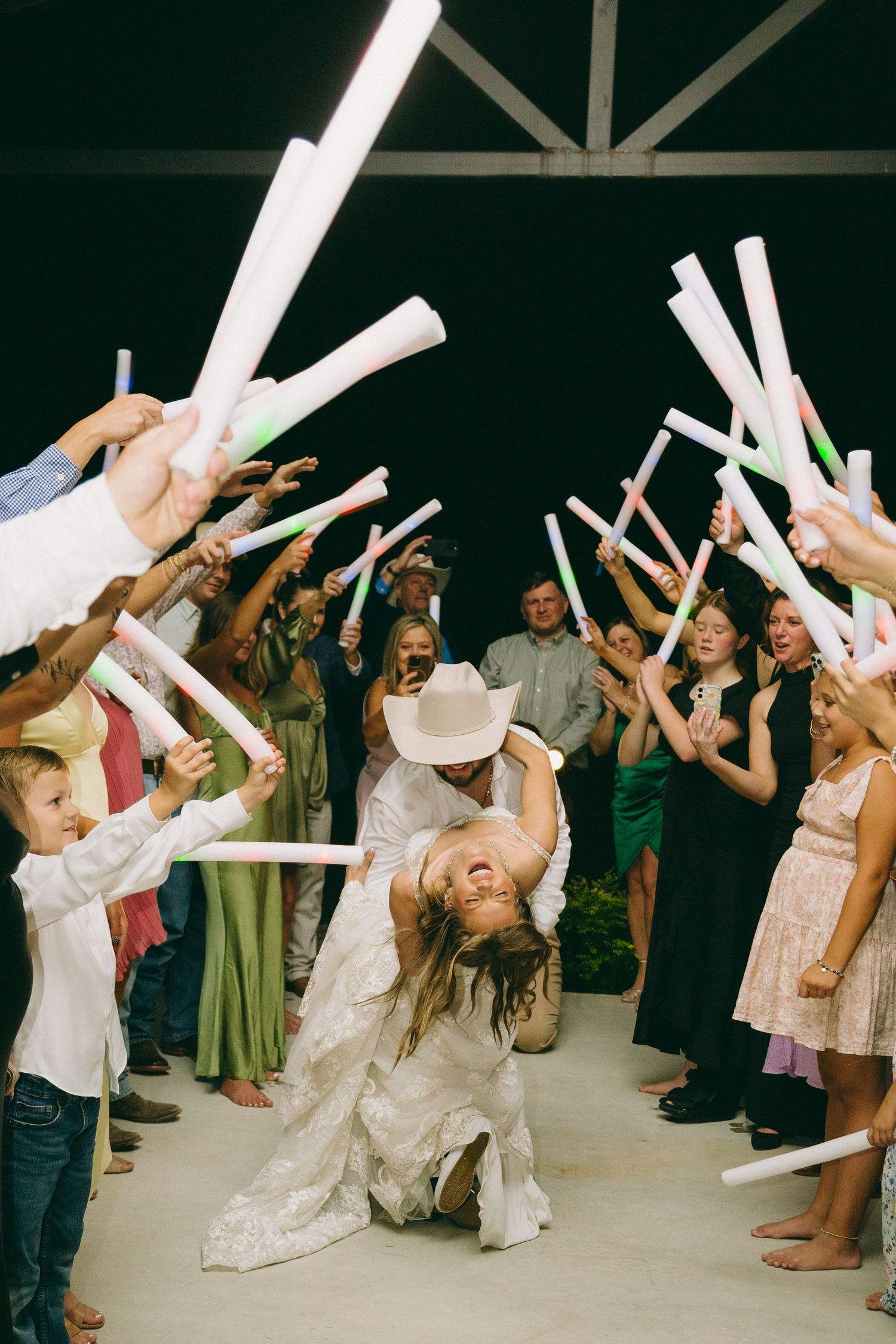 Wedding guests wave white glow sticks as couple dips in celebration on dance floor.