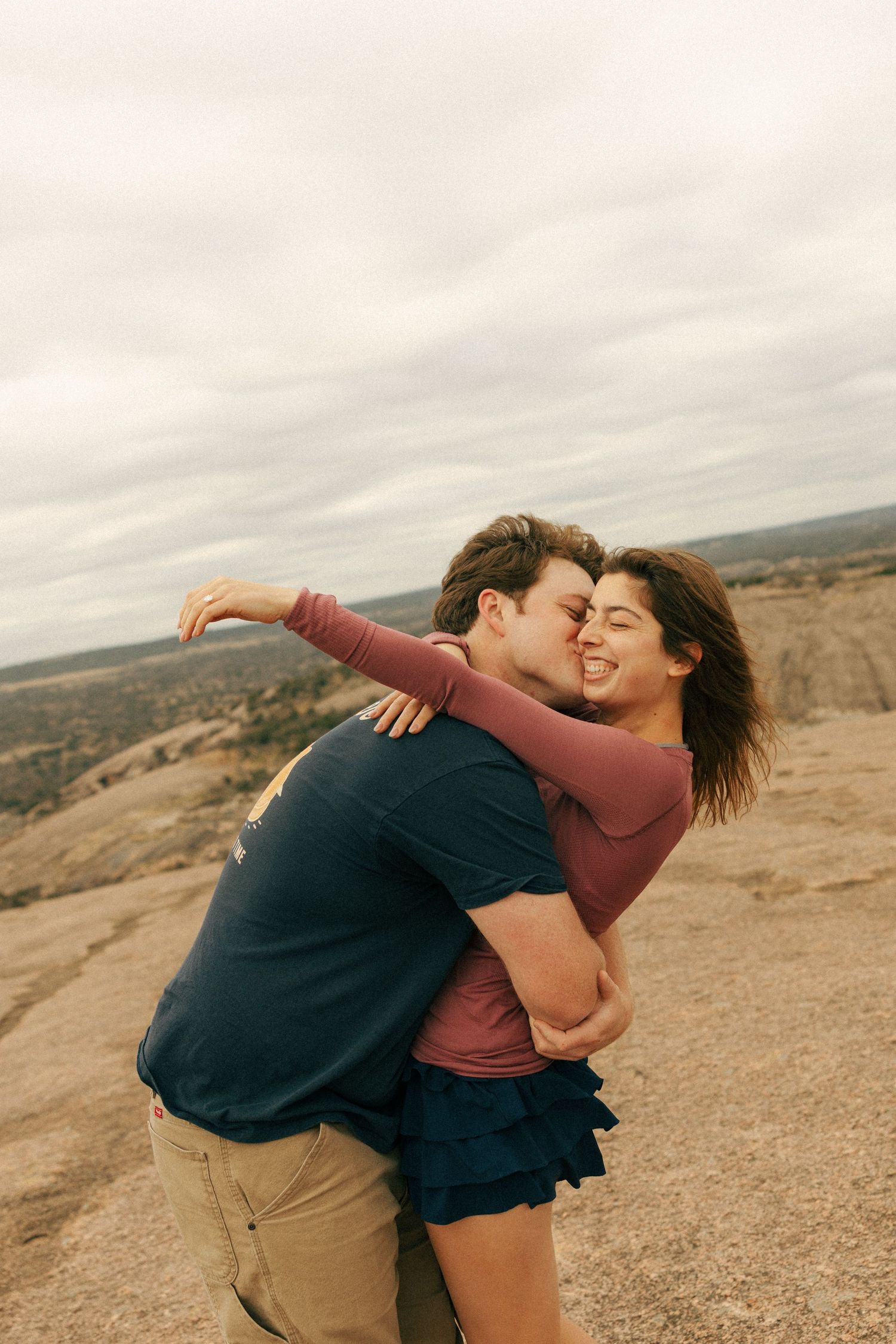 A couple sharing a joyful embrace on a desert cliff with overcast skies.
