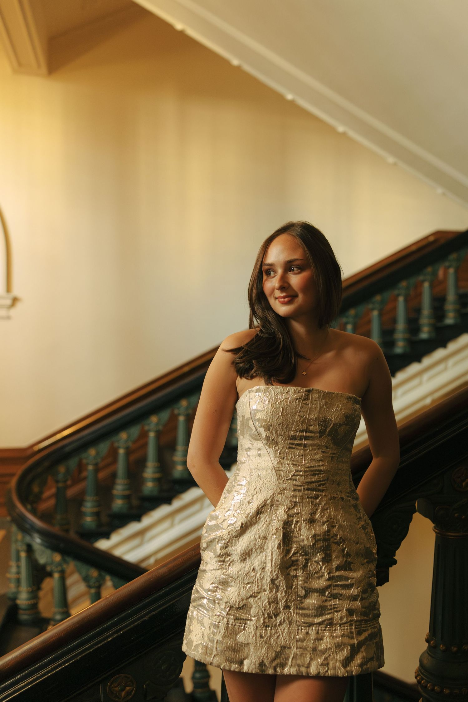 A woman in a textured cream dress poses against an elegant staircase indoors.