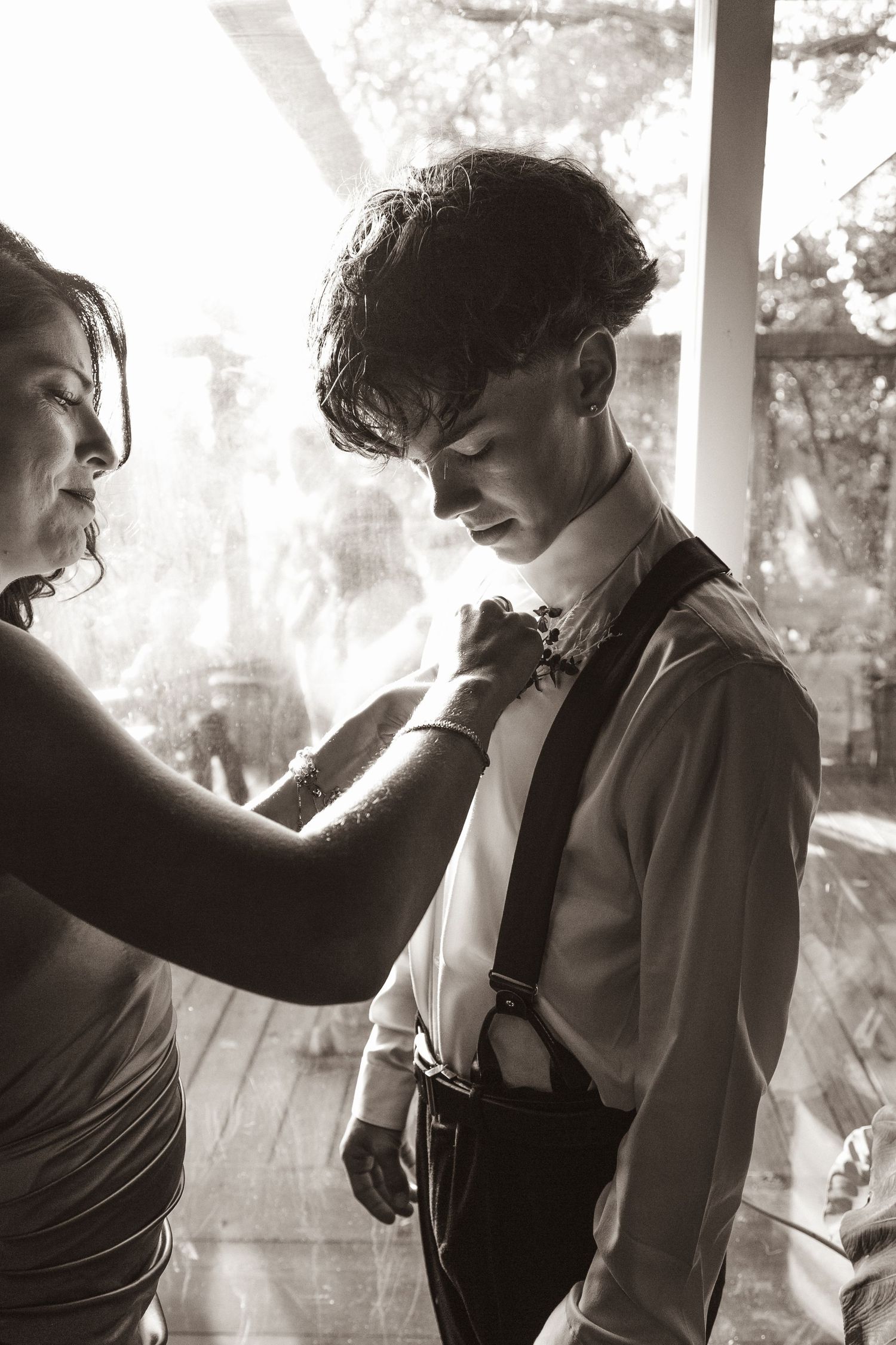 Soft natural light illuminates a scene of a woman placing a boutonnière on a young man before a wedding ceremony.