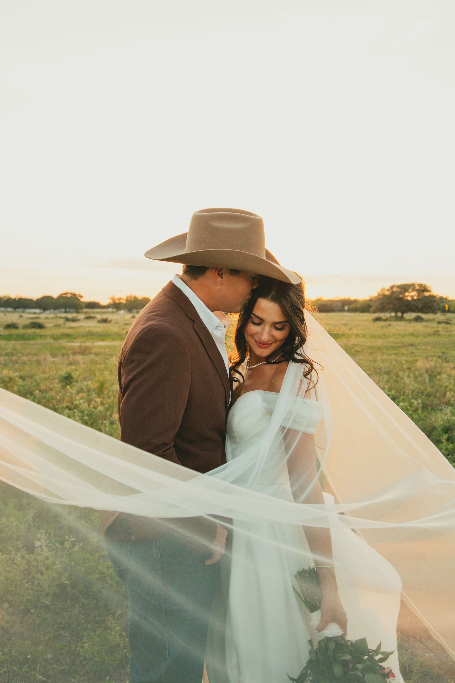 Romantic couple with flowing veil in field during golden hour wedding photography session.