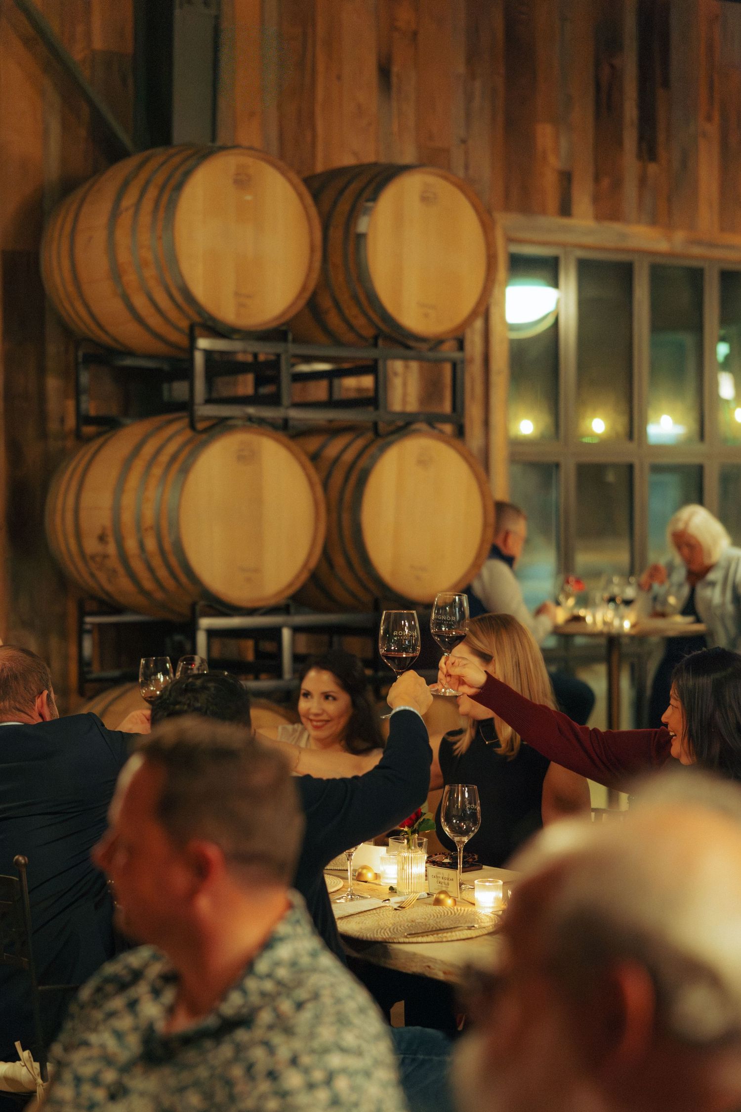 Wine barrels line the walls of a rustic tasting room as guests enjoy an evening gathering.