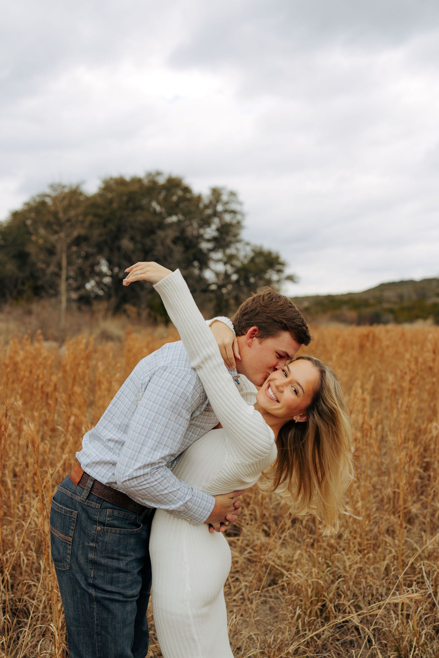 Couple embracing in a rustic field with tall grass and cloudy skies.