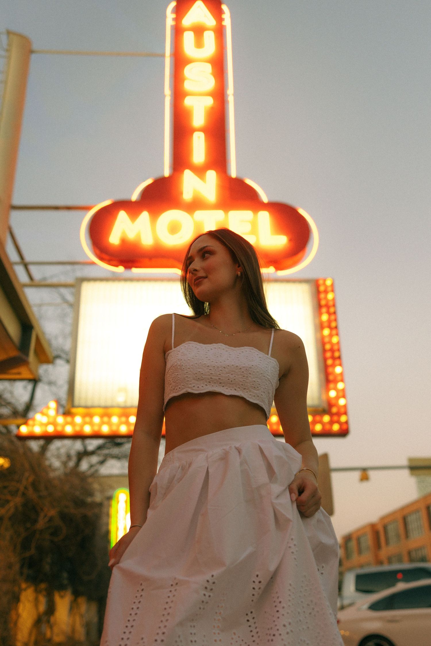 Woman in white dress against illuminated vintage motel sign at dusk.