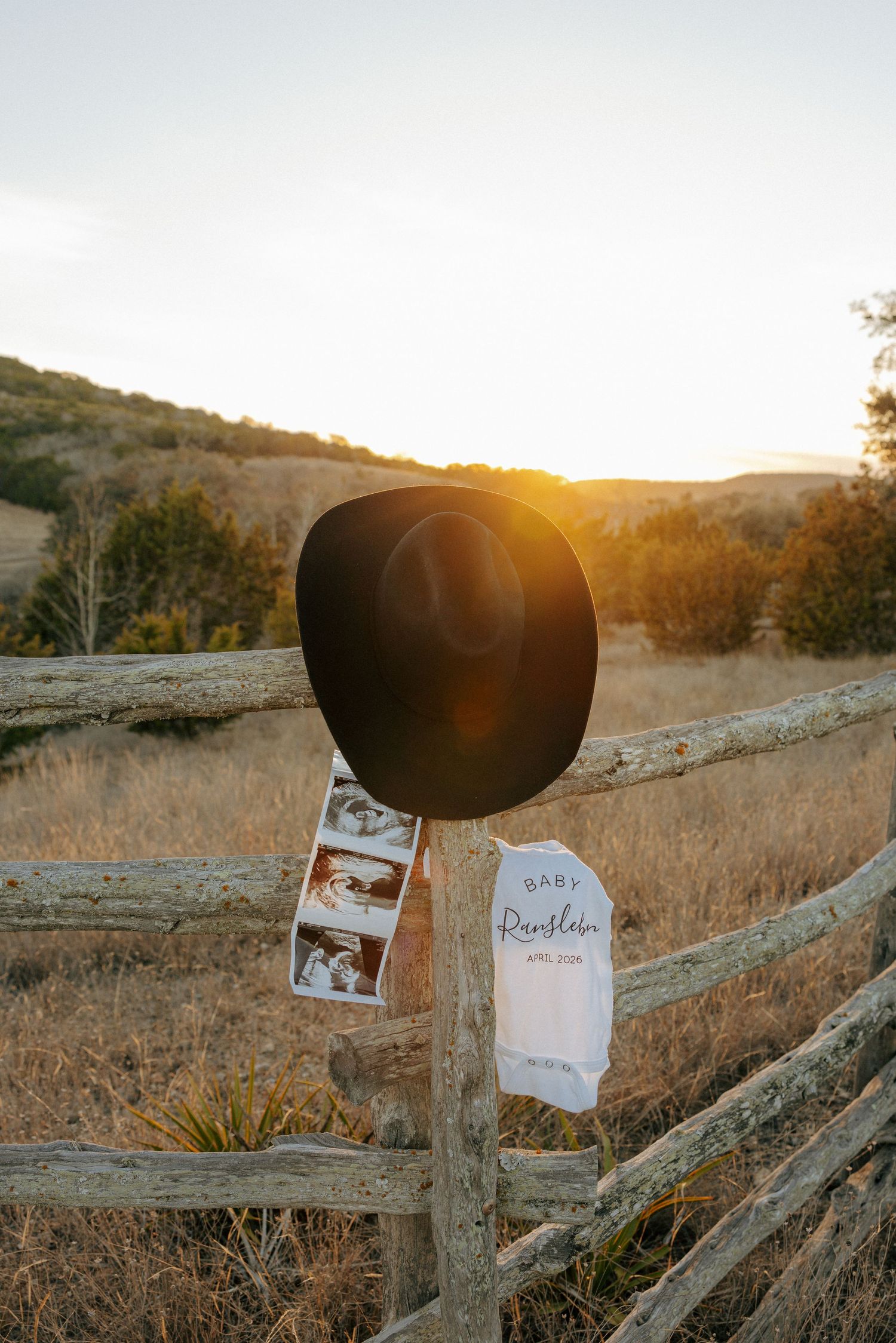 Sonogram photos, a cowboy hat, and a baby onesie displayed on a wooden fence at sunset.