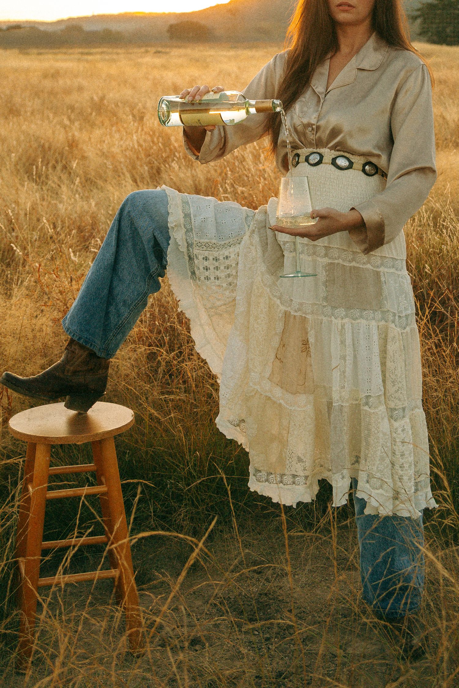 A woman in a flowing white lace skirt pours a glass of wine in a golden field at dusk.