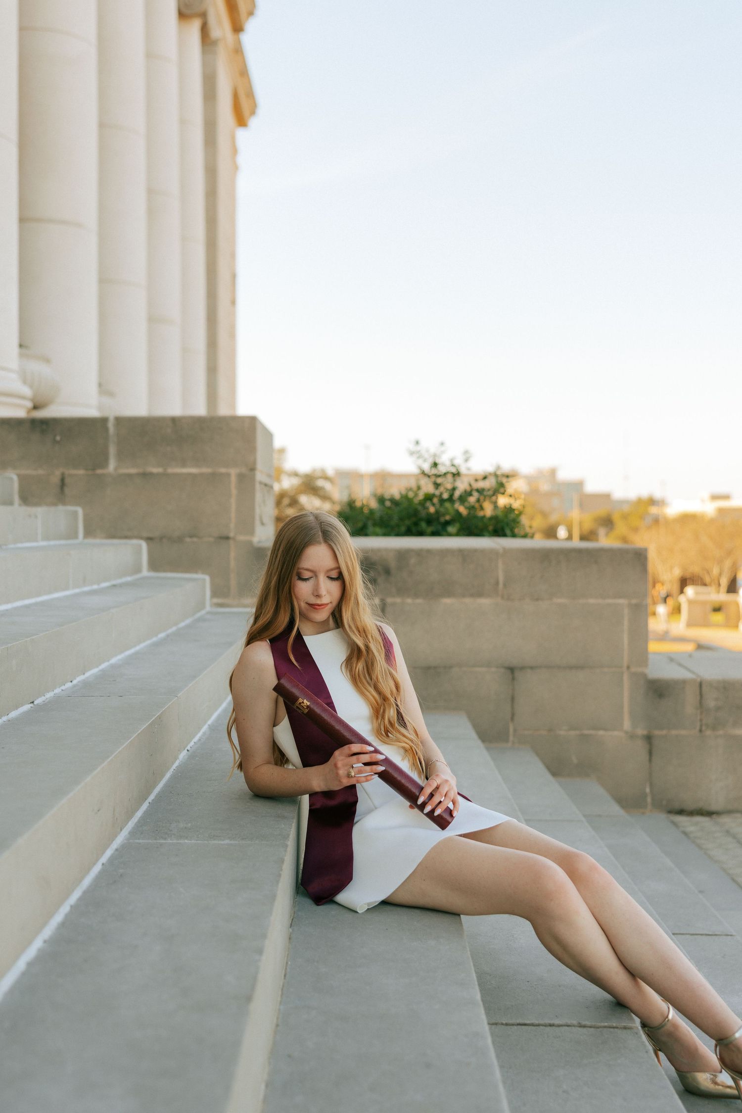 A graduate in a burgundy dress holding a diploma reclines on concrete steps near white columns at sunset.