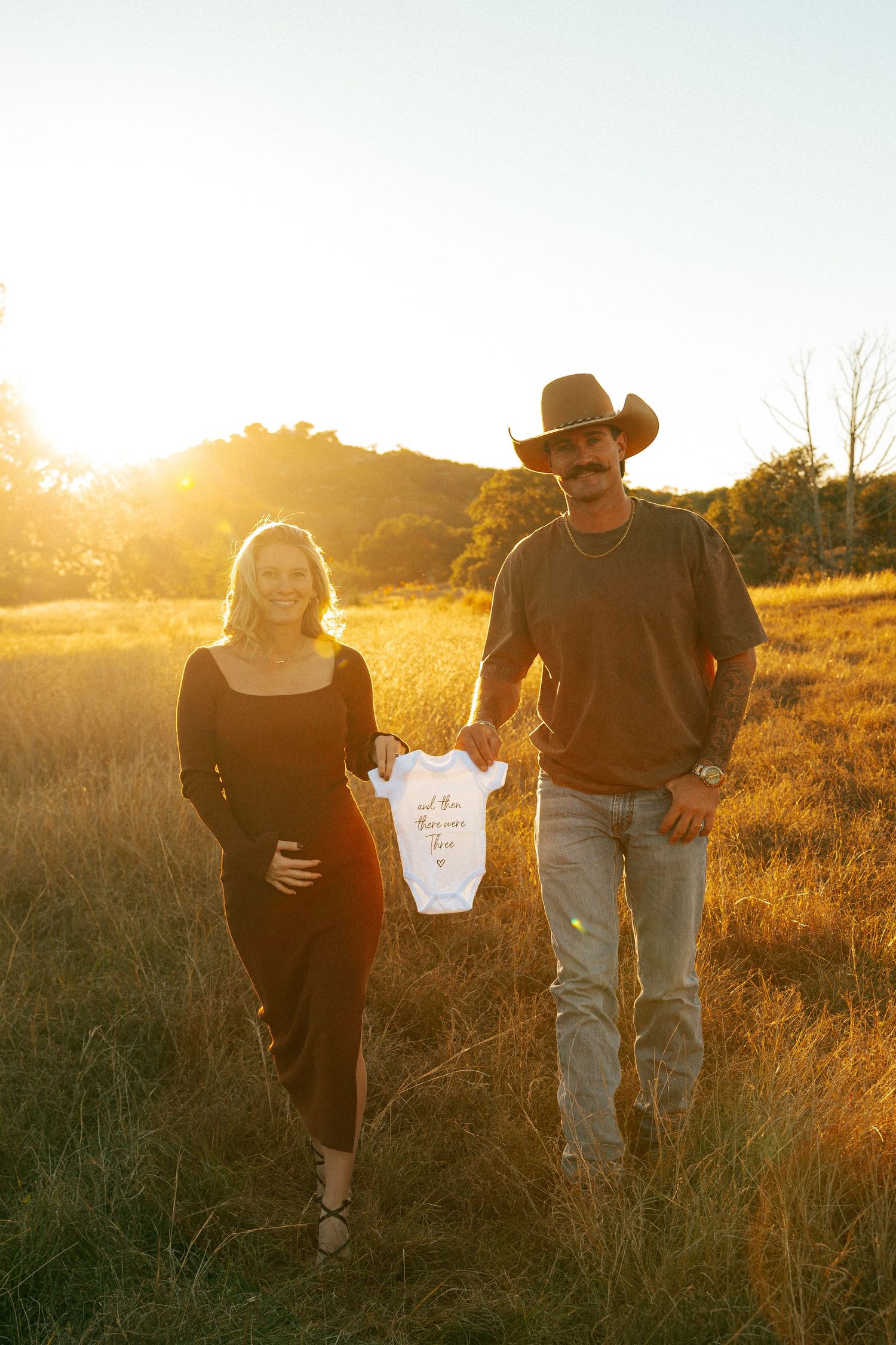 A couple walks through a sunlit field holding a baby onesie during sunset.