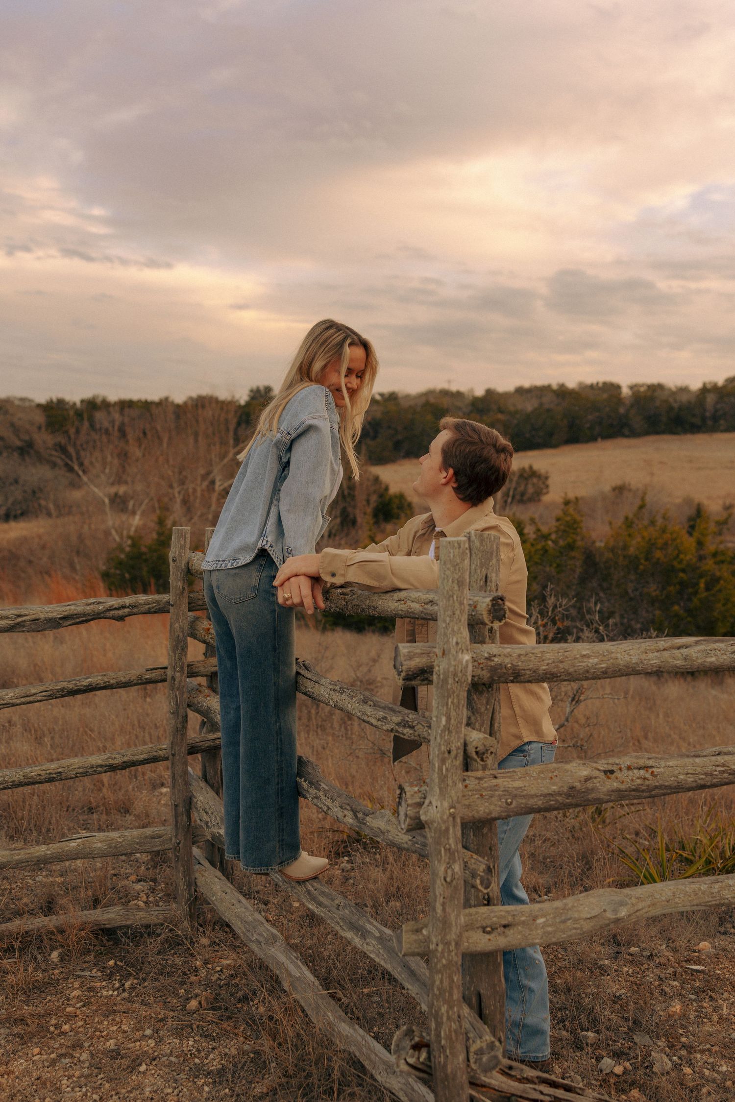 A couple shares a playful interaction by a weathered fence in a rural setting at sunset.
