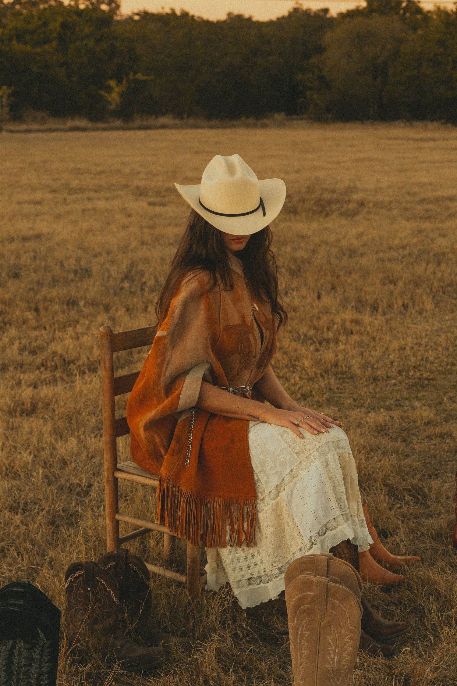 Person in white dress and cowboy hat sitting on wooden chair in golden field surrounded by western boots.