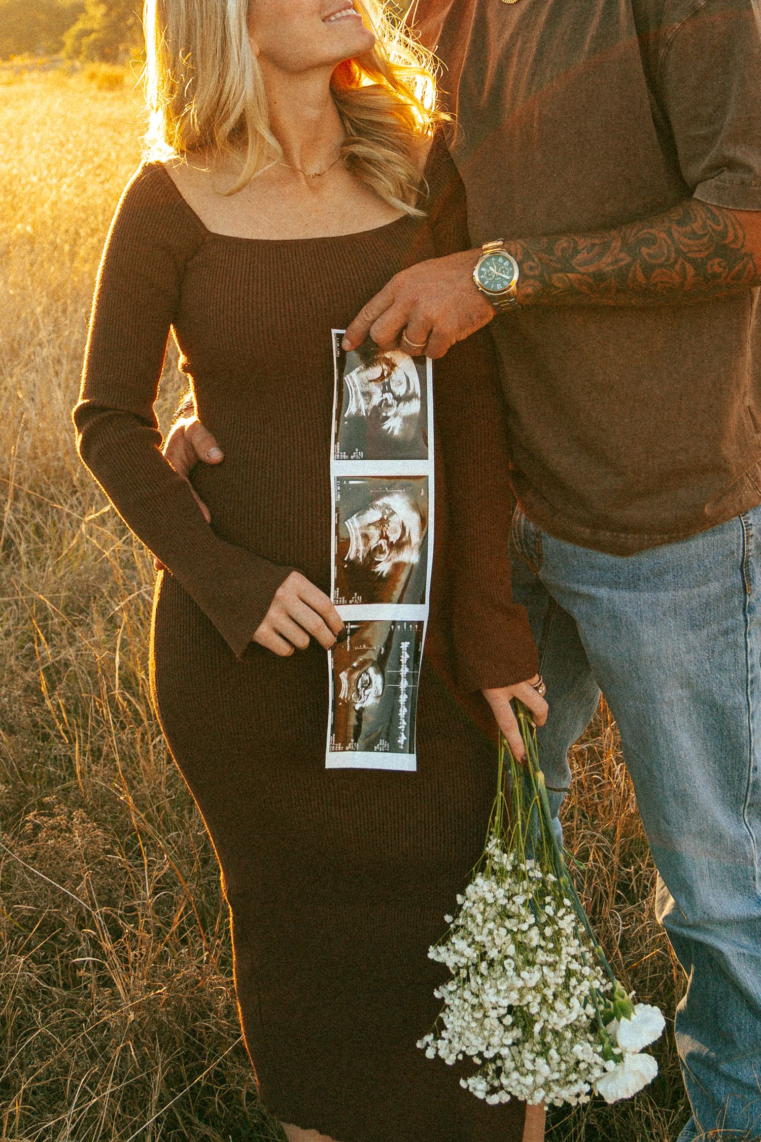 A couple poses with sonogram in a sun-drenched field at golden hour.