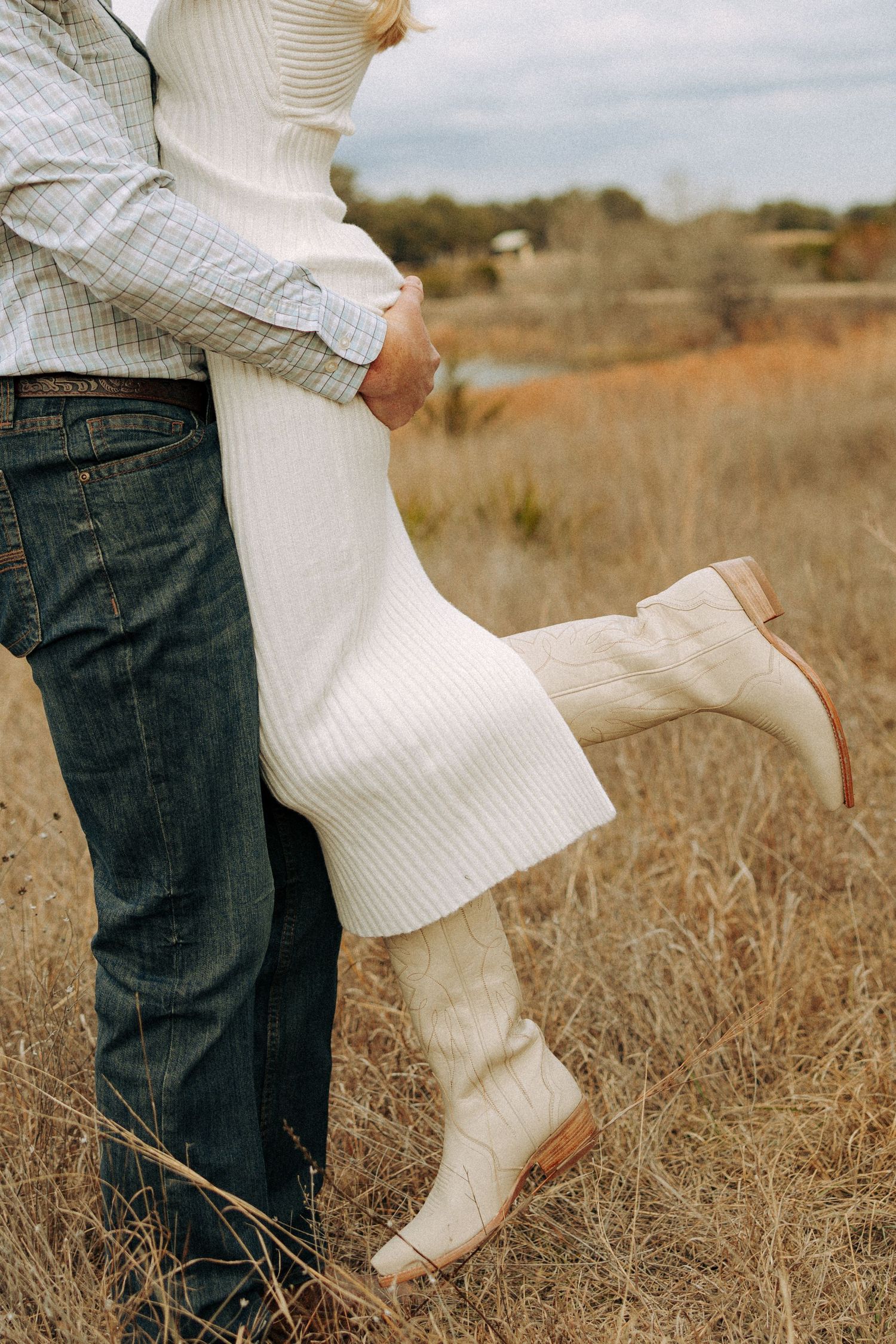 A couple embraces in a field, with focus on her white dress and boots.