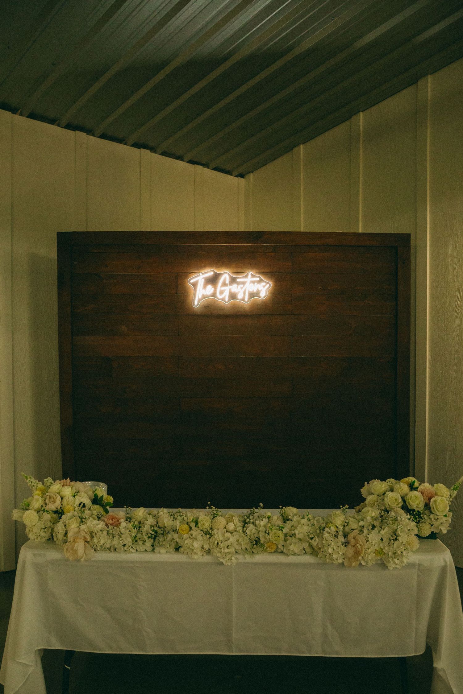 Wedding reception venue with white floral arrangements and neon sign decor against dark wooden walls.