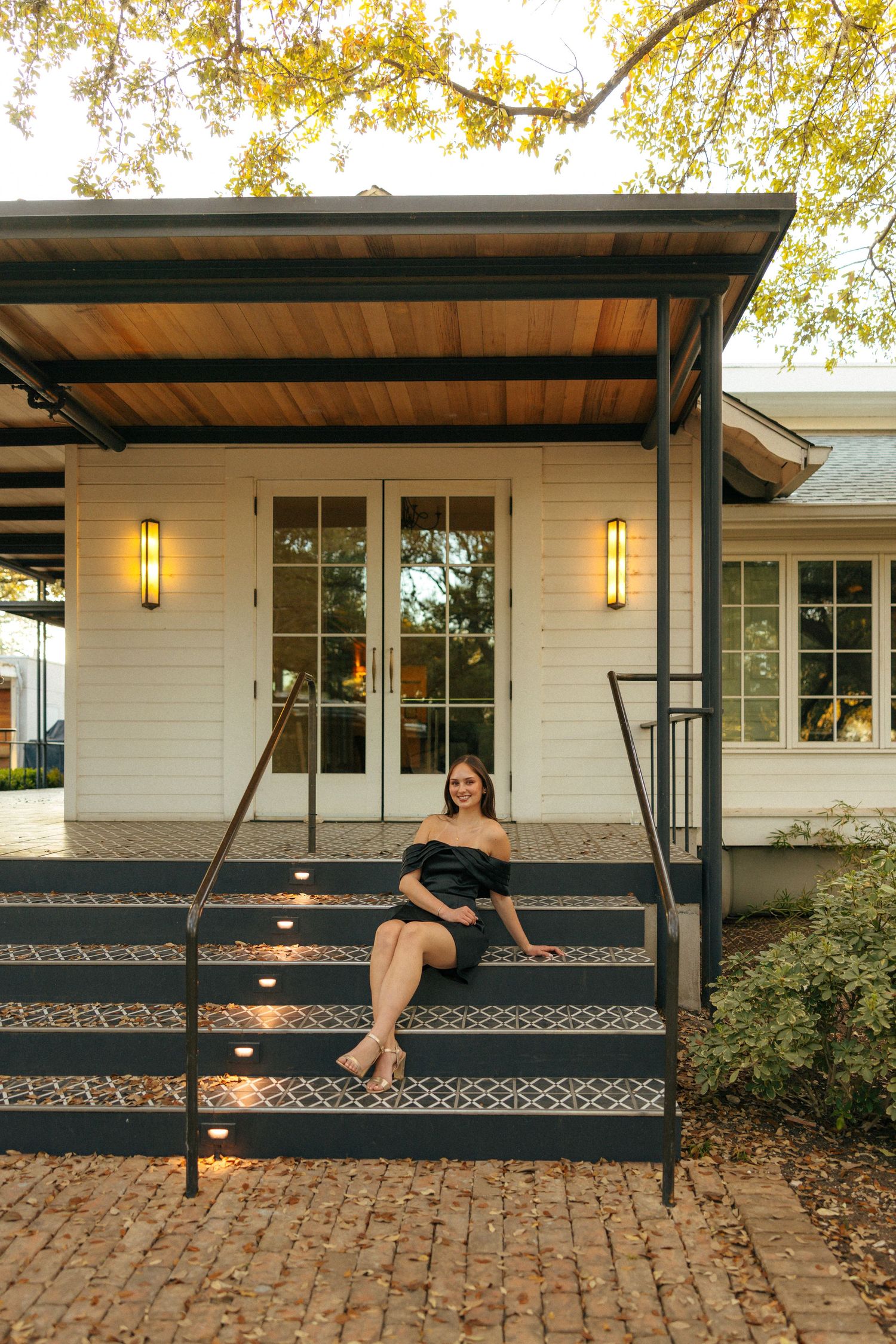 Woman poses on stairs in front of a white farmhouse with a covered porch and lantern lighting at dusk.