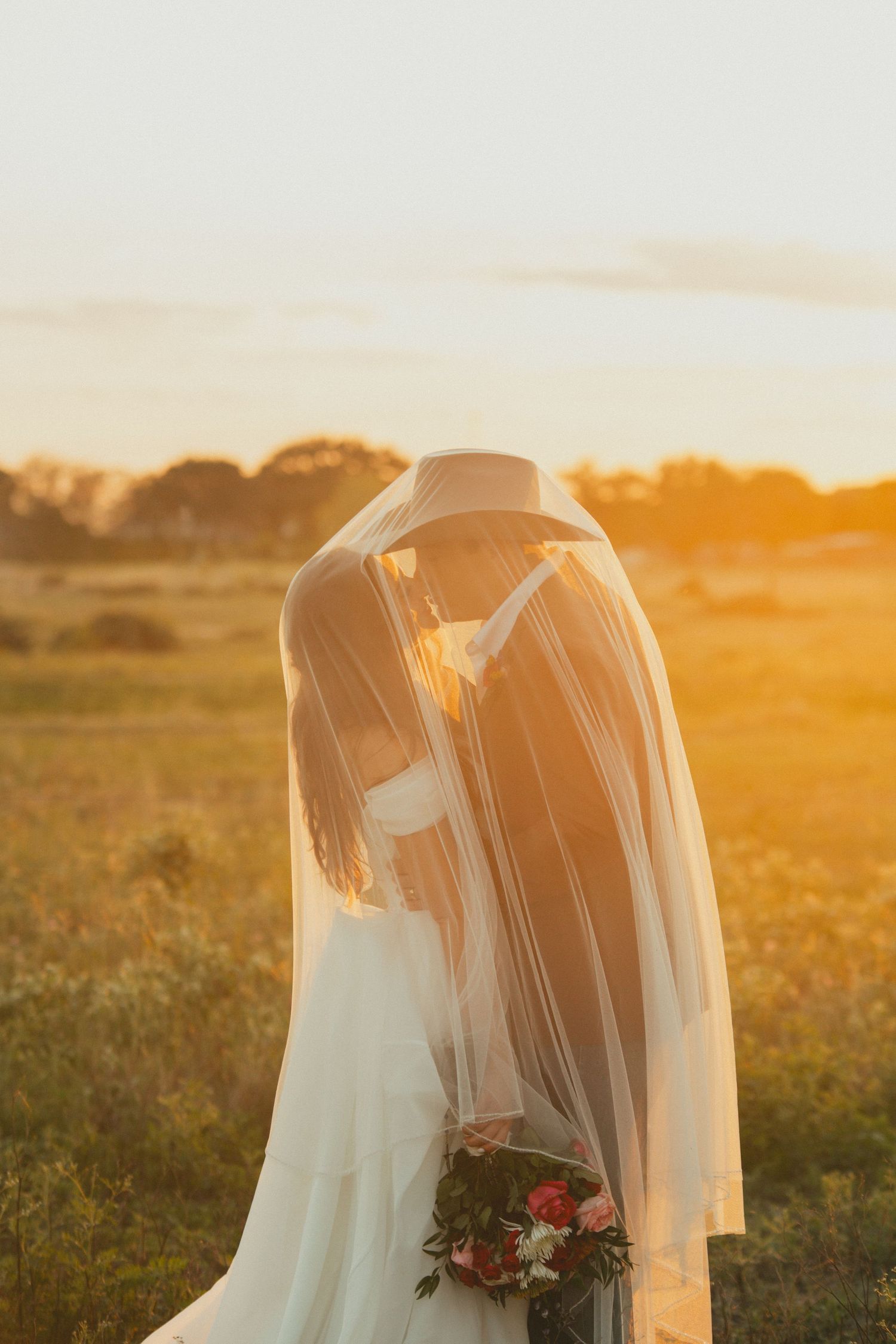 Couple shares an intimate moment together under a white veil in golden sunset light during wedding photoshoot.